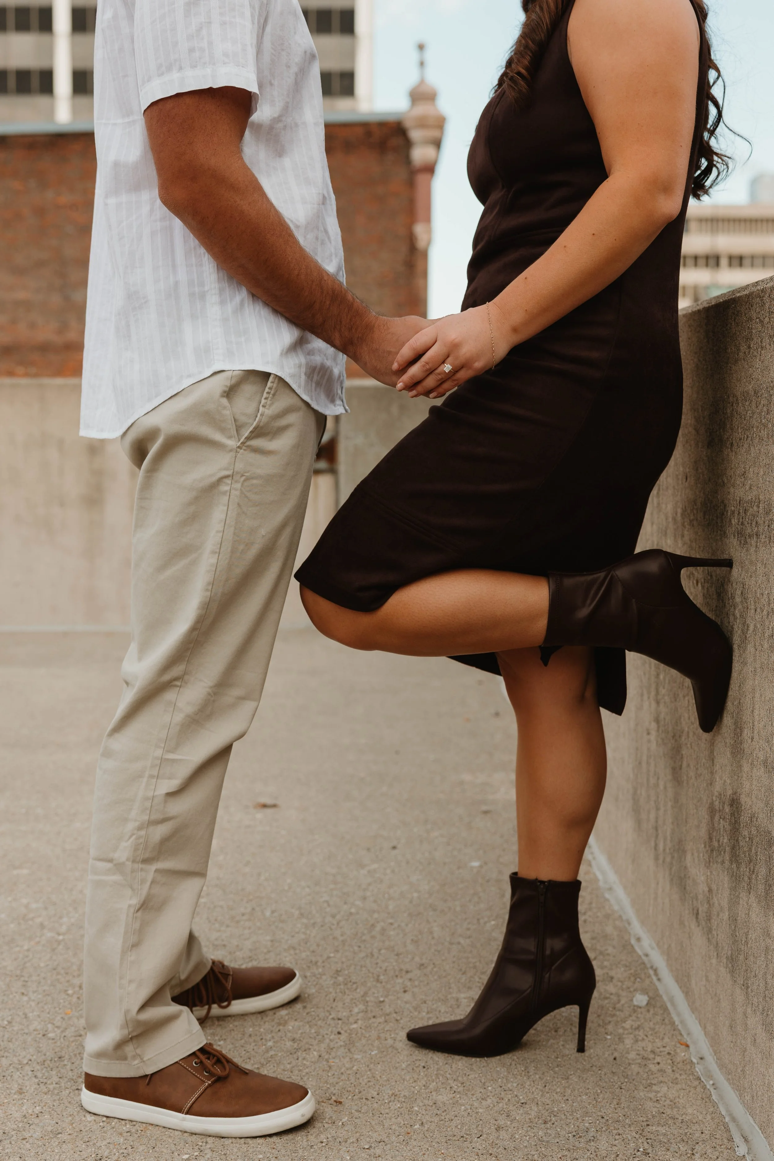 A couple holding hands on a rooftop, with the woman kneeling and wearing black high-heeled boots and a black dress, and the man standing in beige pants and brown shoes.