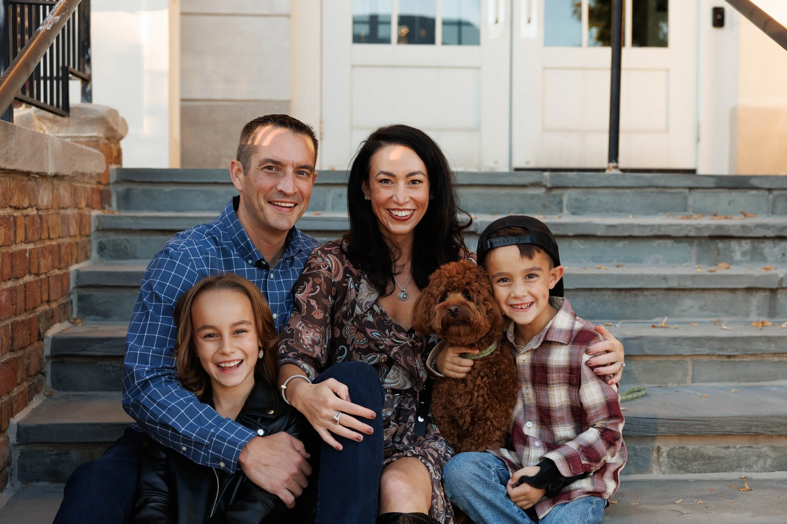 A family sitting on outdoor steps with a dog, smiling at the camera.