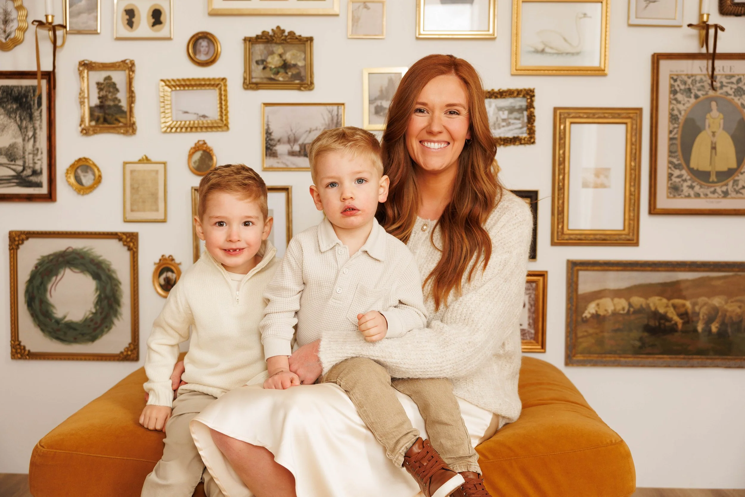 A woman with long red hair smiling while sitting on a yellow couch with two young boys, all dressed in light sweaters, in front of a wall decorated with numerous gold-framed pictures and artwork.