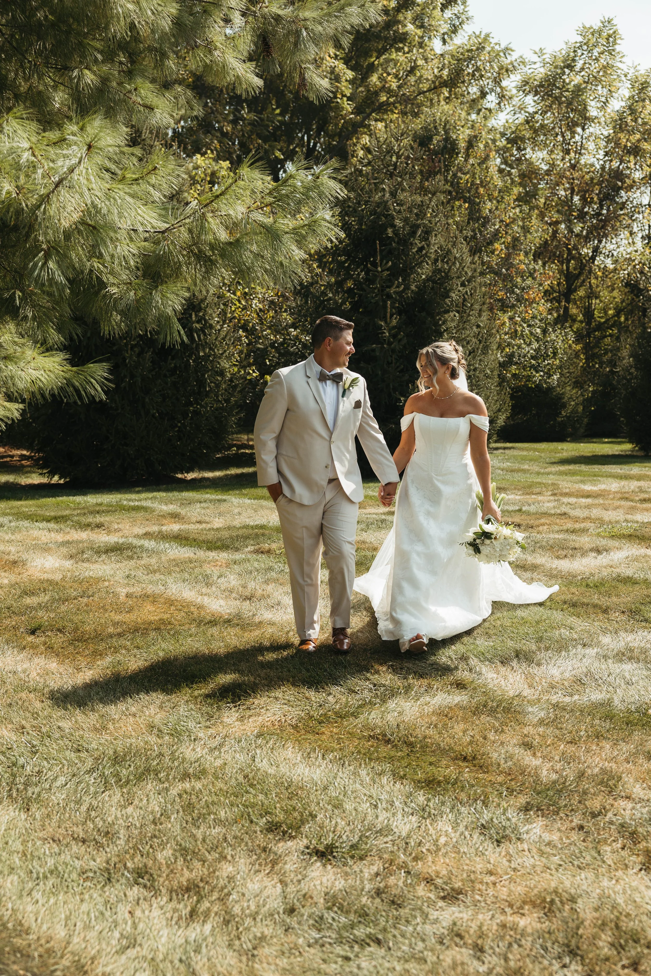 A happy bride and groom walking hand in hand in a park-like setting on their wedding day, surrounded by green trees and grass, with sunlight filtering through the foliage.