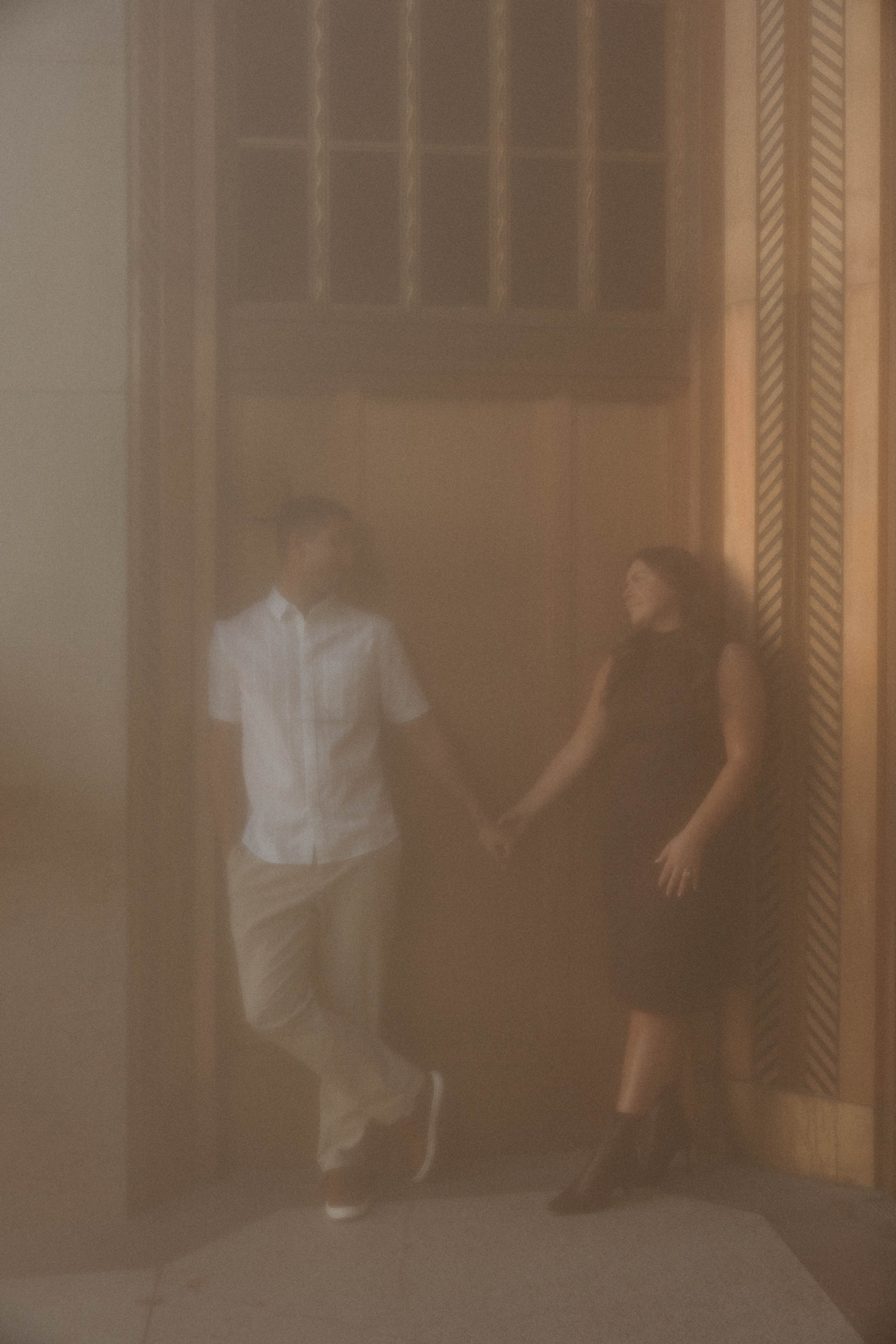Two people holding hands and leaning against a doorway in a softly lit room with wooden paneling.