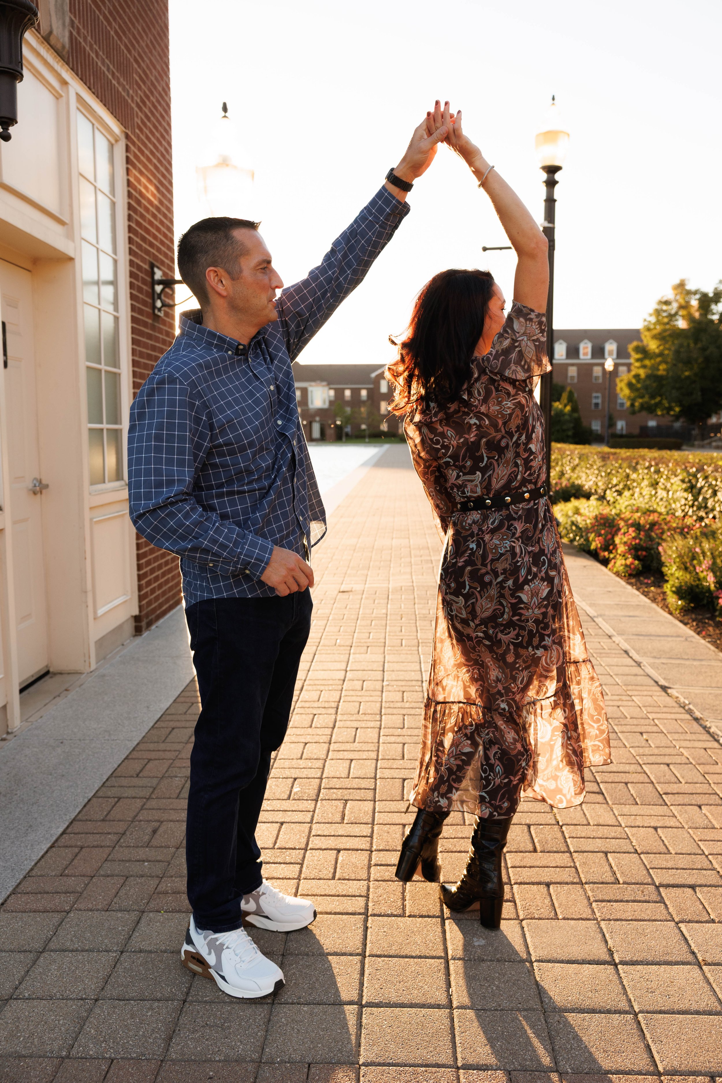 A man and a woman dancing together outdoors during sunset. The man wears a checkered shirt, black pants, and white sneakers. The woman wears a floral dress and black high-heeled boots. They hold hands in a dance pose, with the woman's arms raised abo