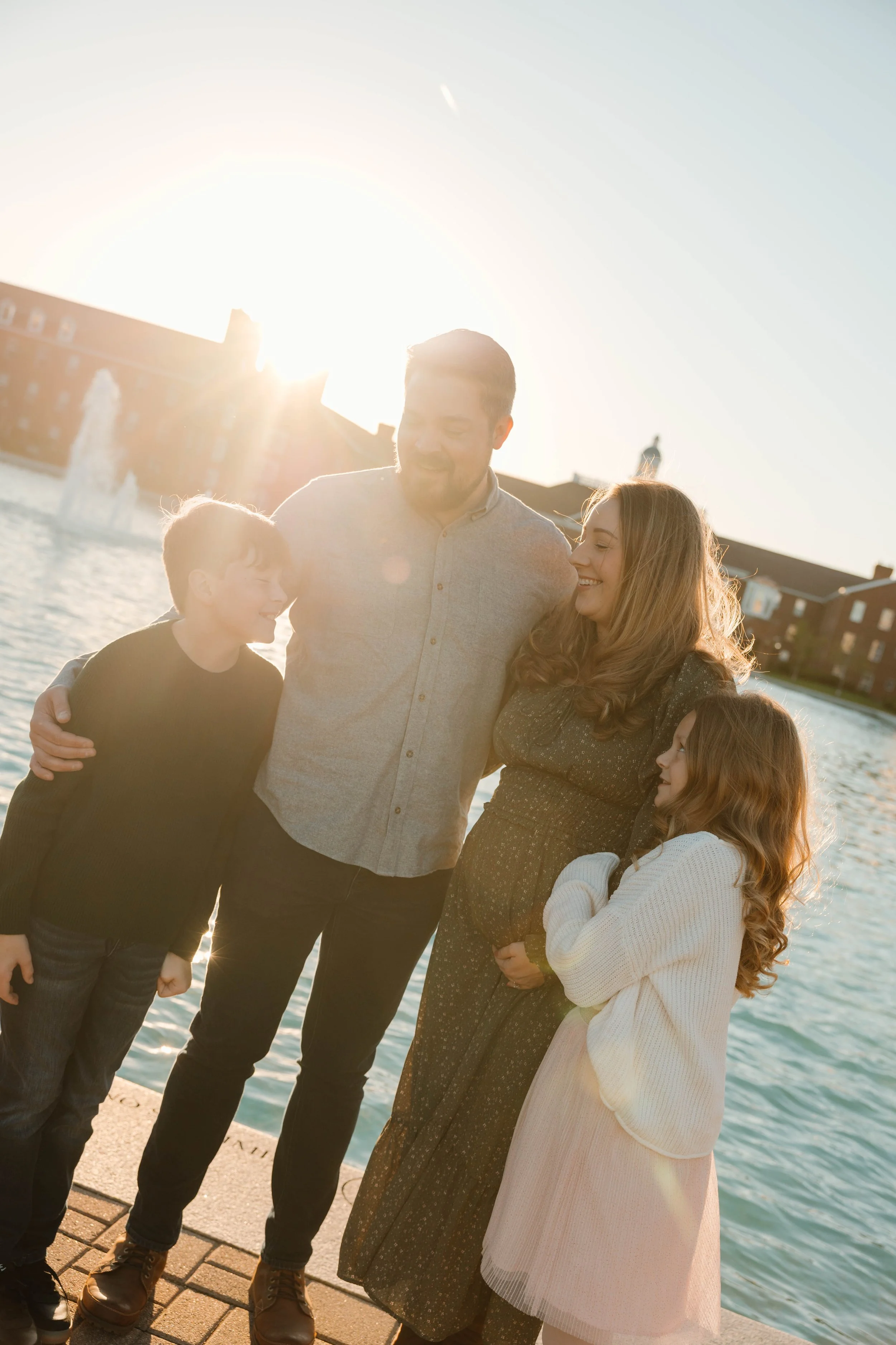 Family of four standing by a fountain near water, smiling and enjoying the sunshine.