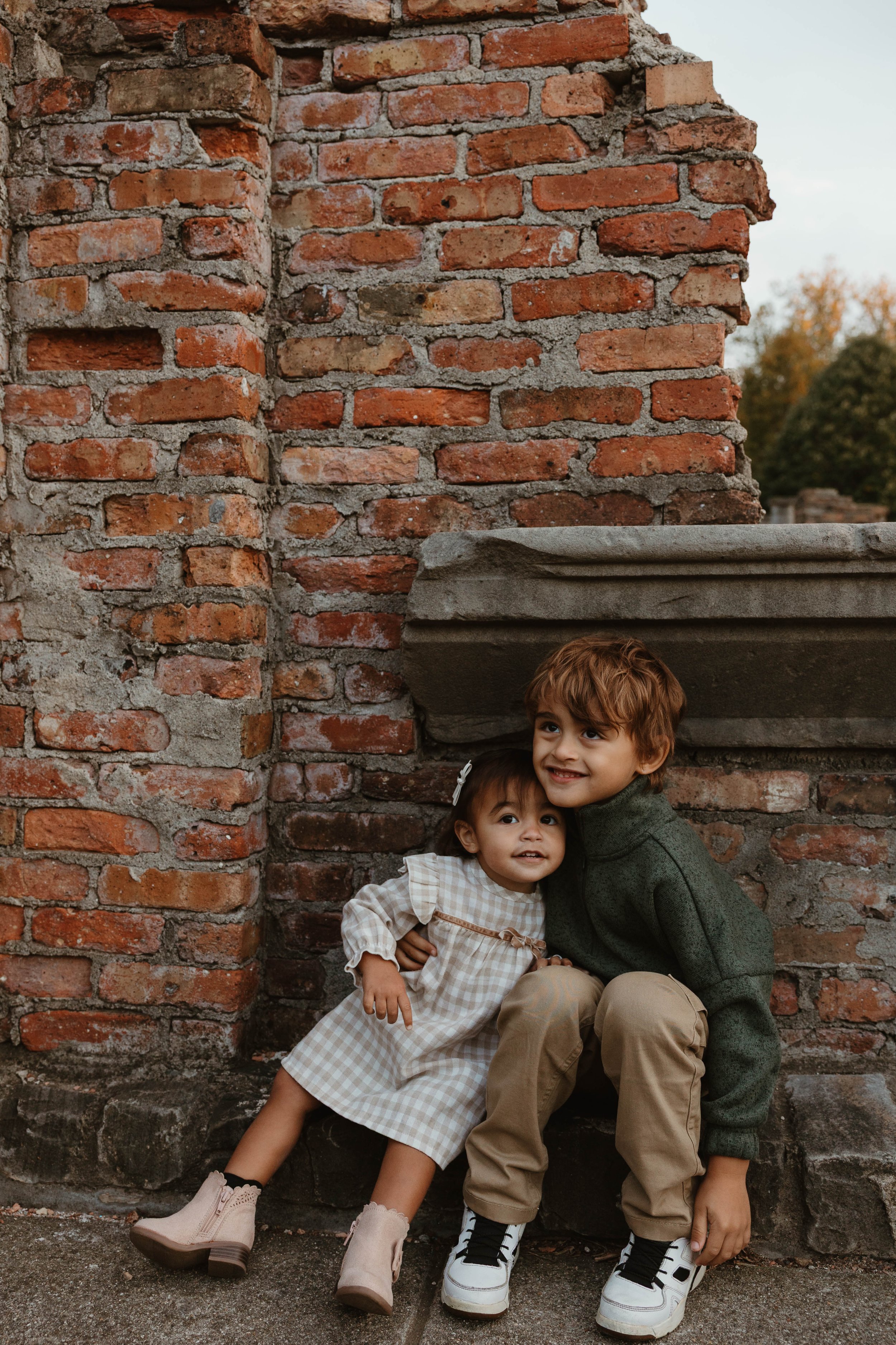 Two children, a girl and a boy, are sitting against a brick wall outdoors, smiling and hugging each other.