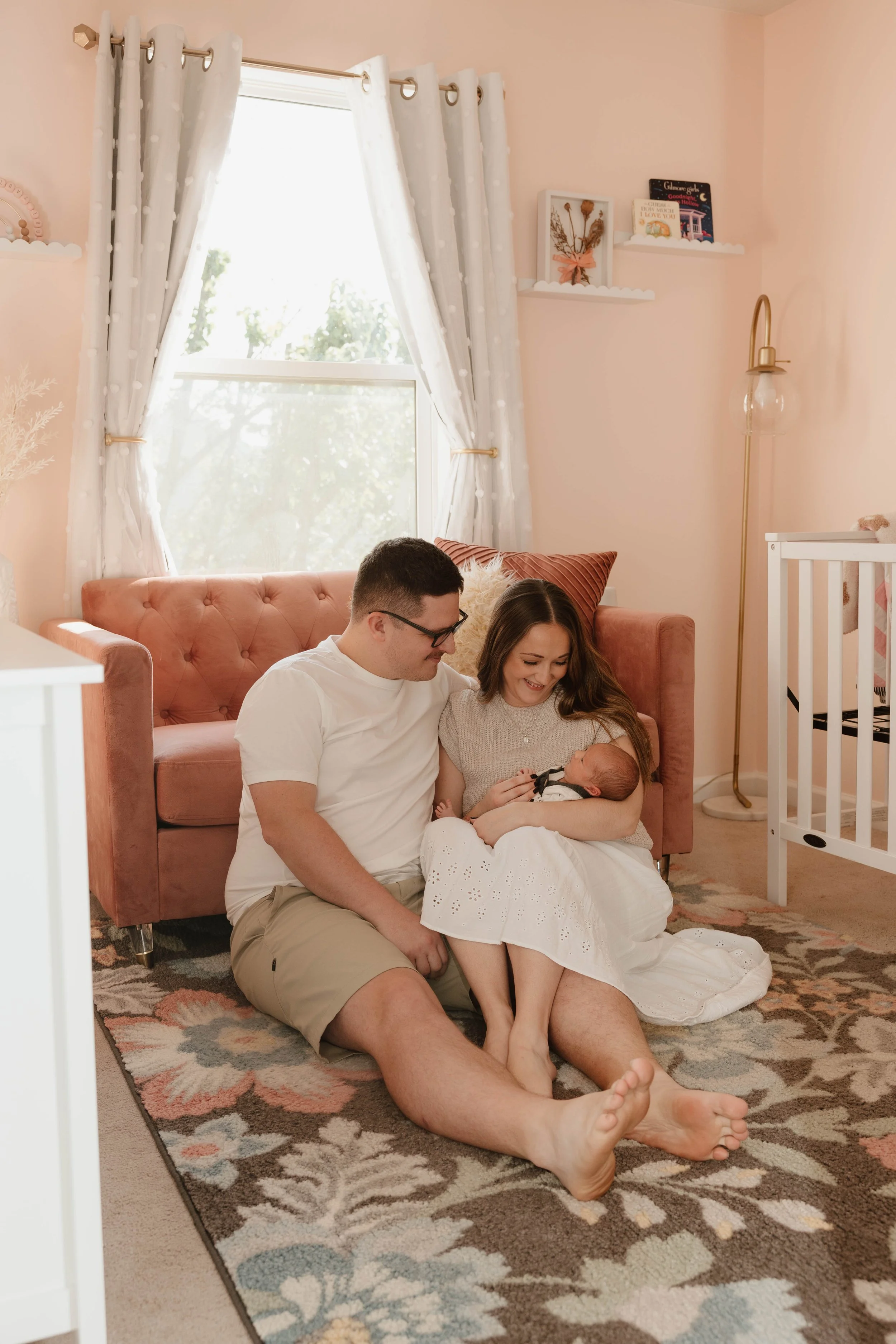 A family of three sitting on the carpeted floor of a nursery, with a woman holding a newborn baby girl, and a man sitting beside her, smiling at the baby. The room has pink walls, a pink tufted sofa, white curtains, and a white crib.