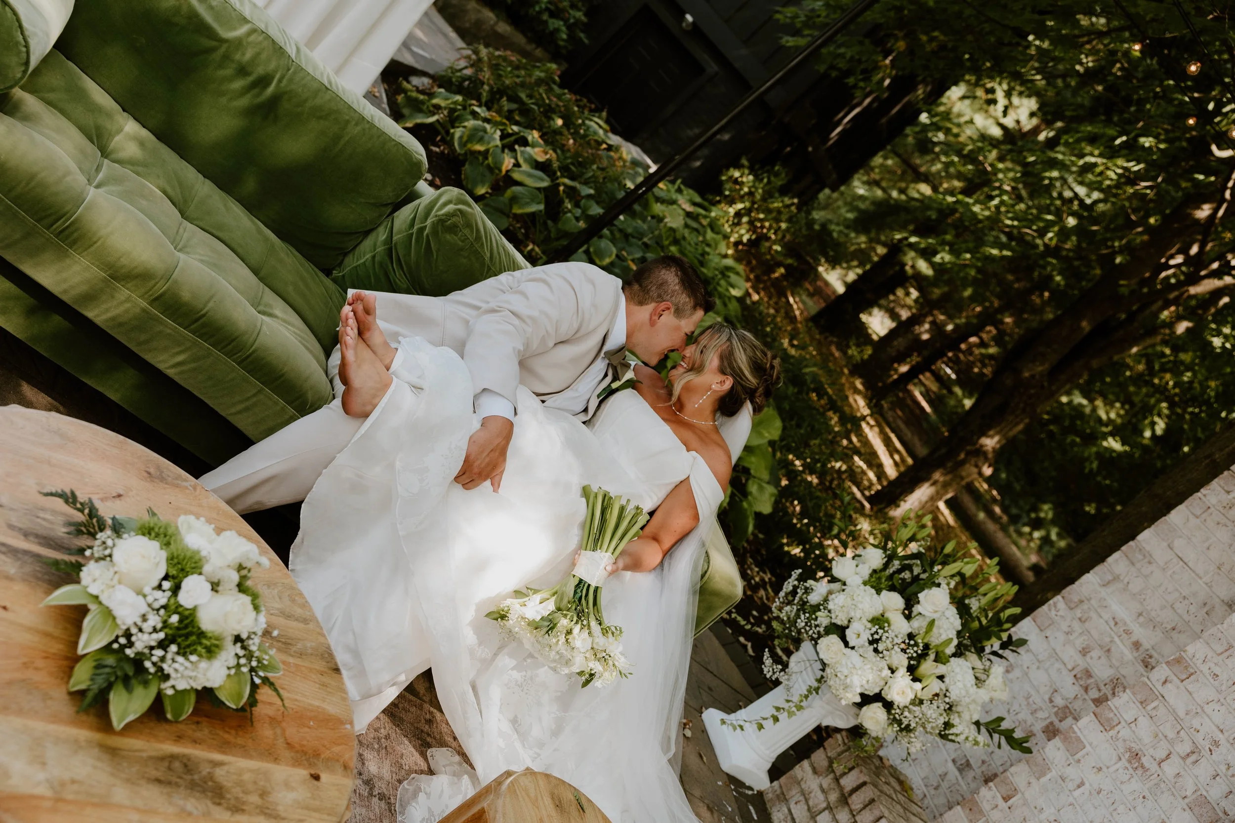 A couple in wedding attire sitting closely on a green sofa outdoors, with the bride holding a bouquet and the groom leaning toward her in a tender moment, surrounded by white floral arrangements and greenery.