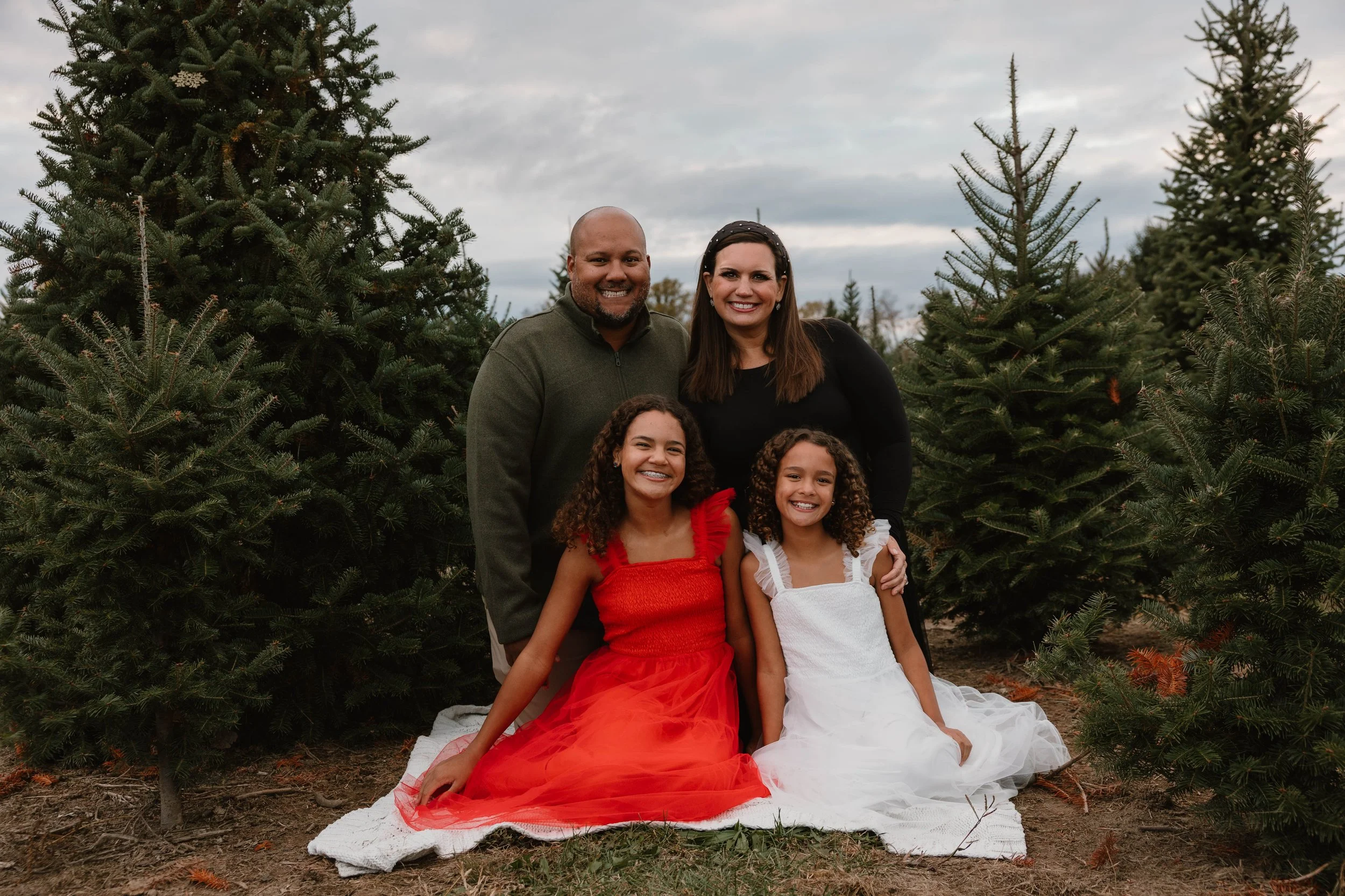 A family of four outdoor during daytime, sitting on a blanket in front of evergreen trees, smiling at the camera.