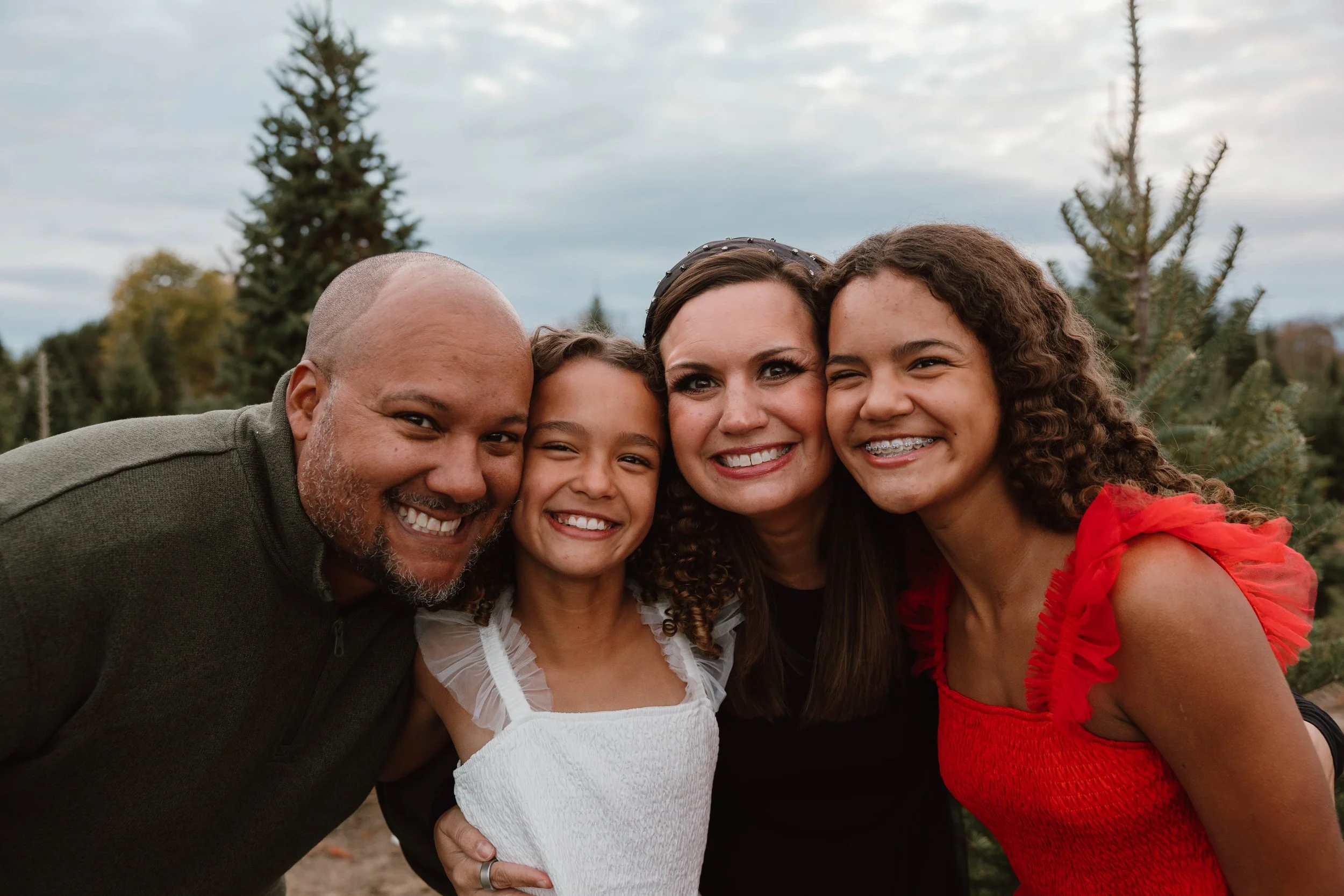 Family of five smiling and posing closely outdoors during daytime, with trees and cloudy sky in the background.