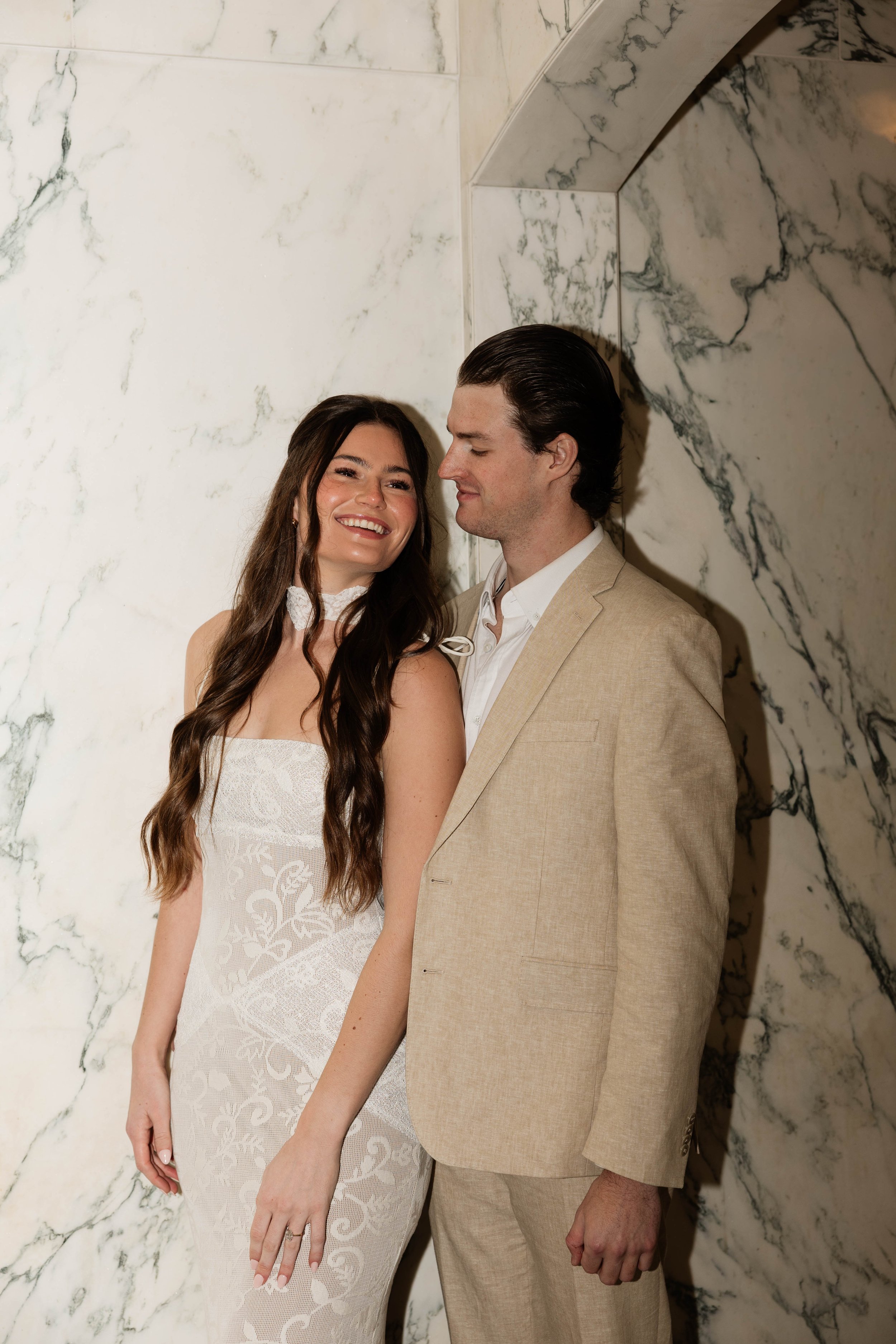 A smiling woman and a man in a beige suit standing close and looking at each other against a white marble wall with dark veins.