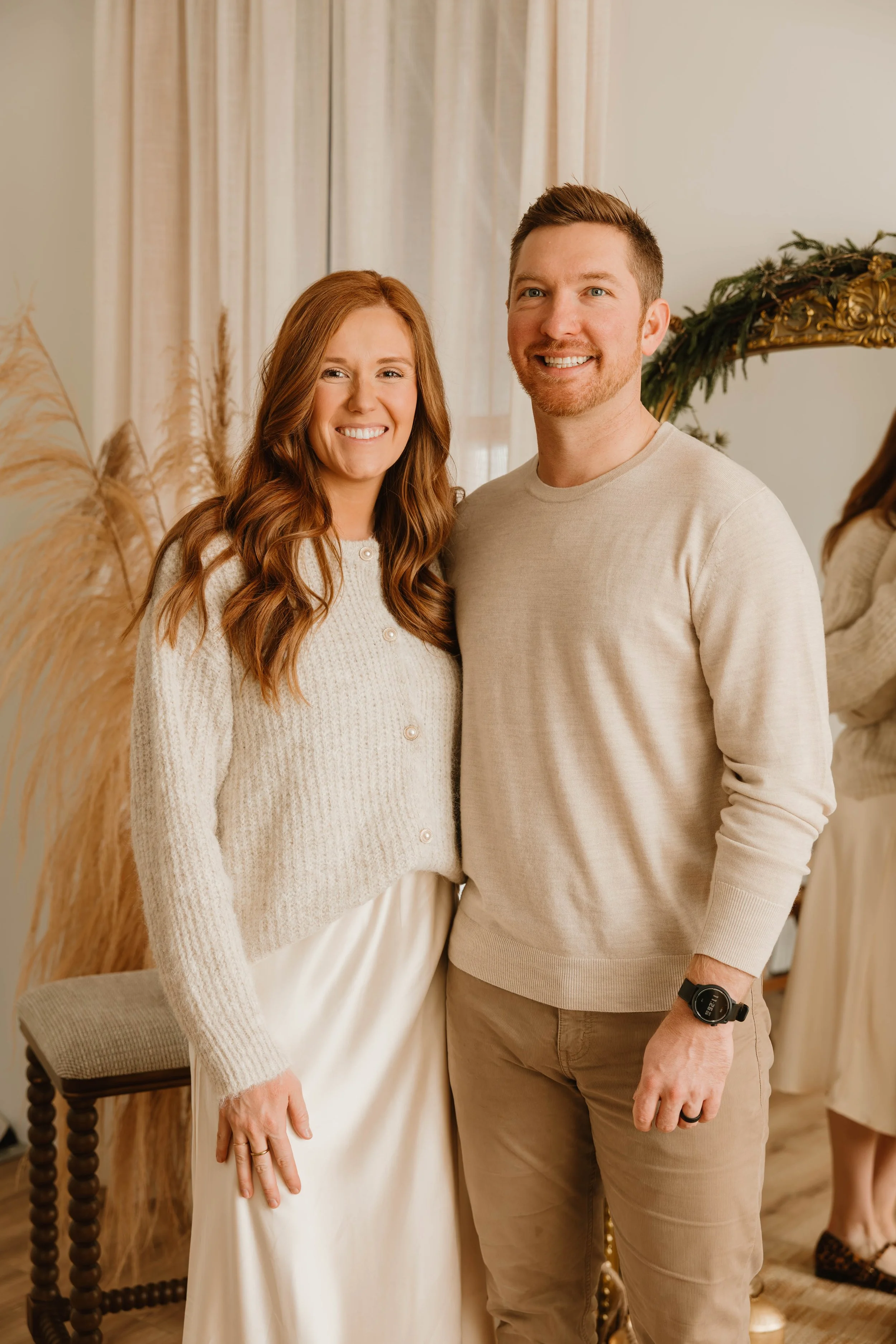 A smiling woman with long wavy red hair and a man with short light brown hair and a beard, standing together in a warmly decorated room with beige curtains and dried pampas grass, posing for a photo.