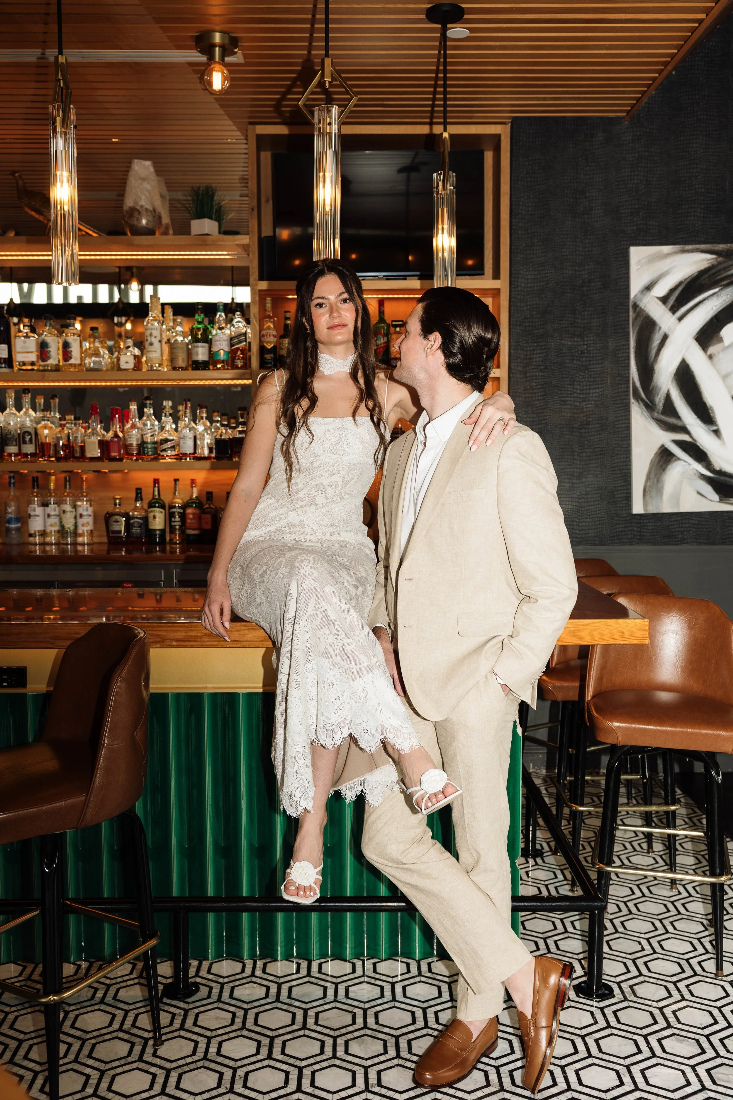 A woman in a white lace dress sitting on a bar counter with a man in a beige suit standing next to her, in a modern bar with bottles on shelves, decorative lighting, and black and white artwork.