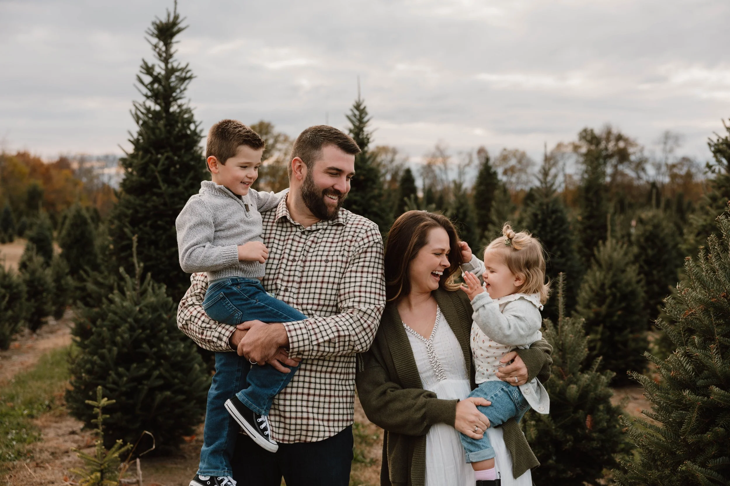 A happy family of four, including a father, mother, young son, and young daughter, enjoying a walk among Christmas trees outdoors during fall, with trees and cloudy sky in the background.
