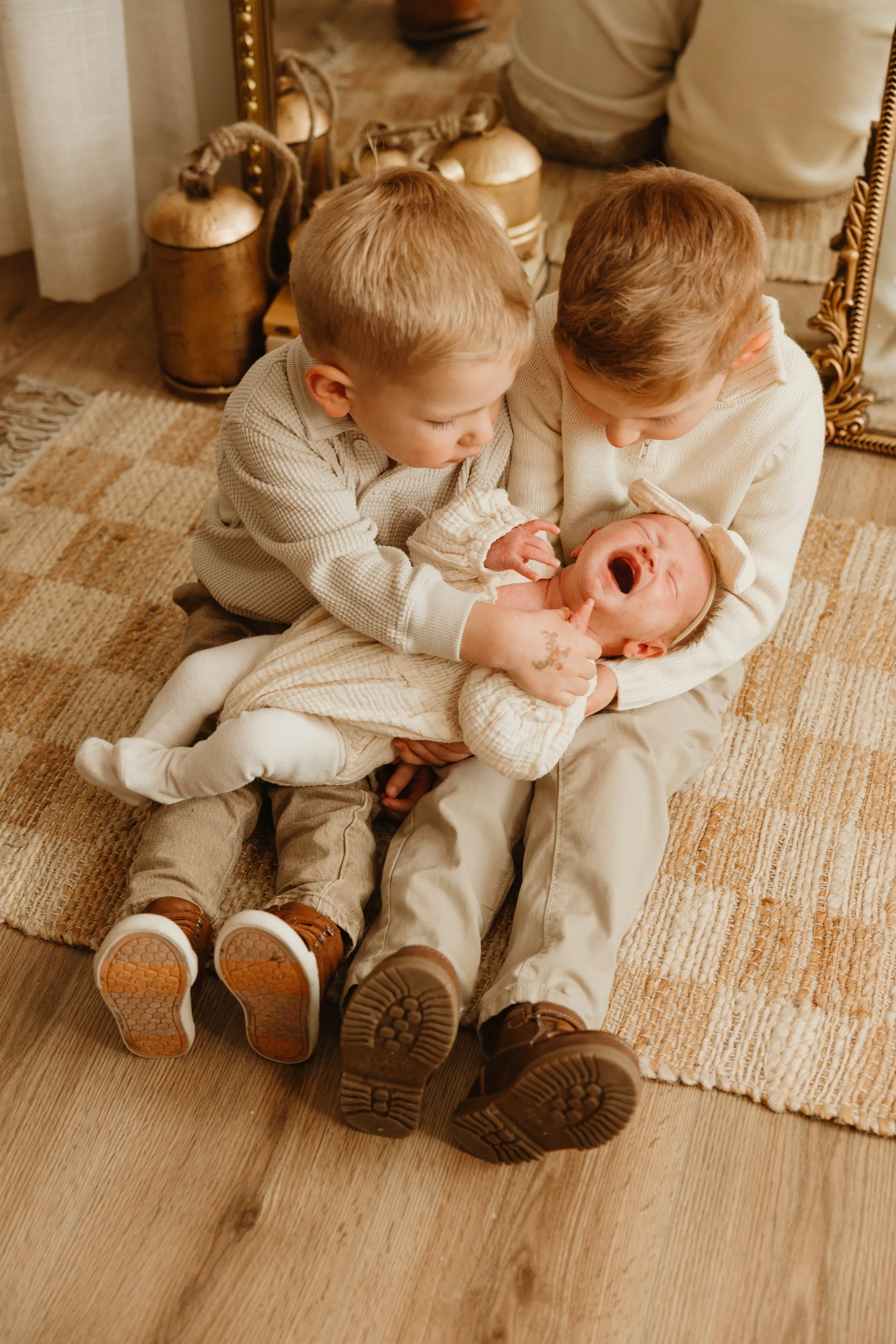 Two young boys holding a crying baby on a wooden floor with a mirror and decorative jars in the background.