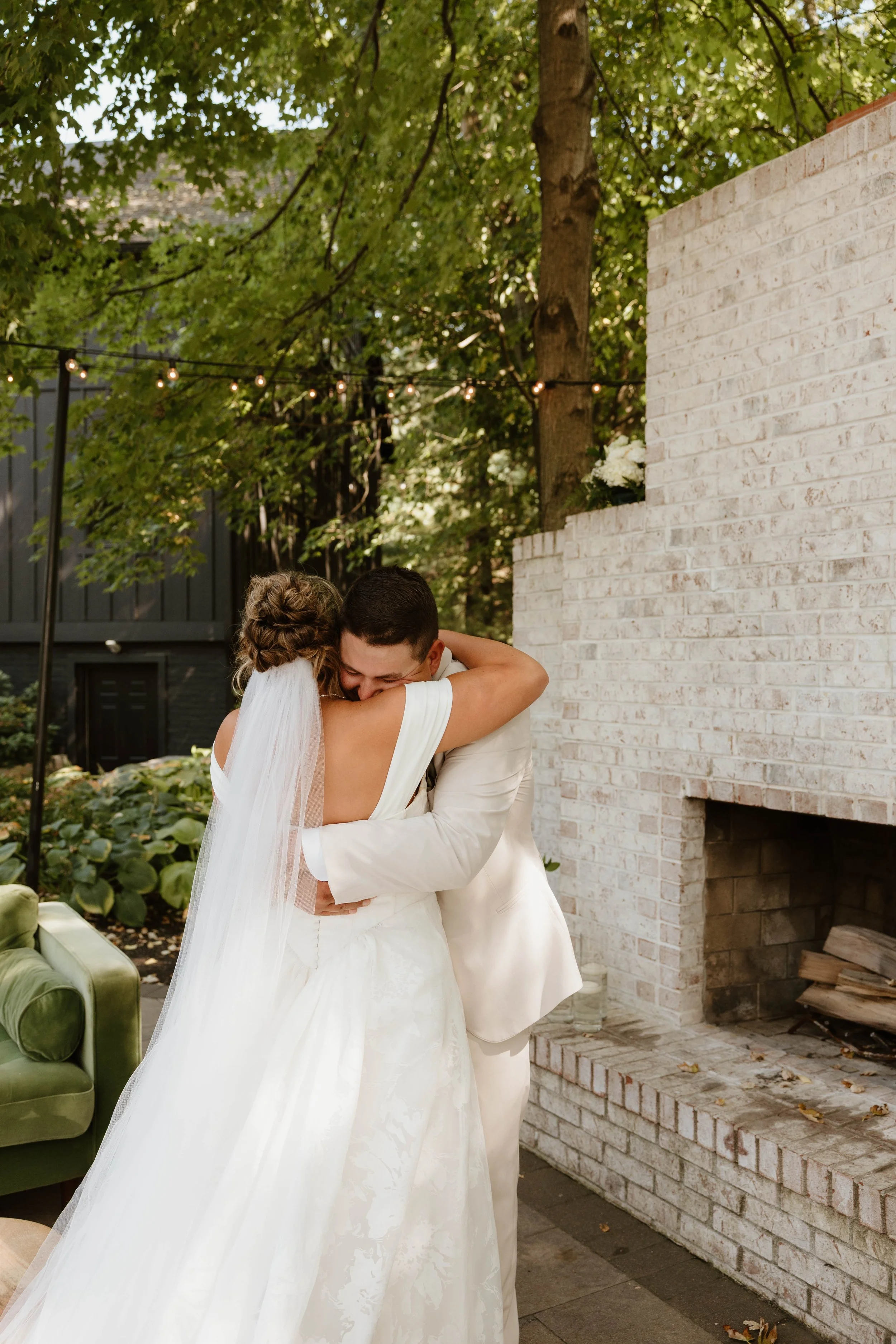 A bride in a white wedding gown and veil hugging a groom in a white suit outdoors near a brick fireplace, under green trees and string lights.