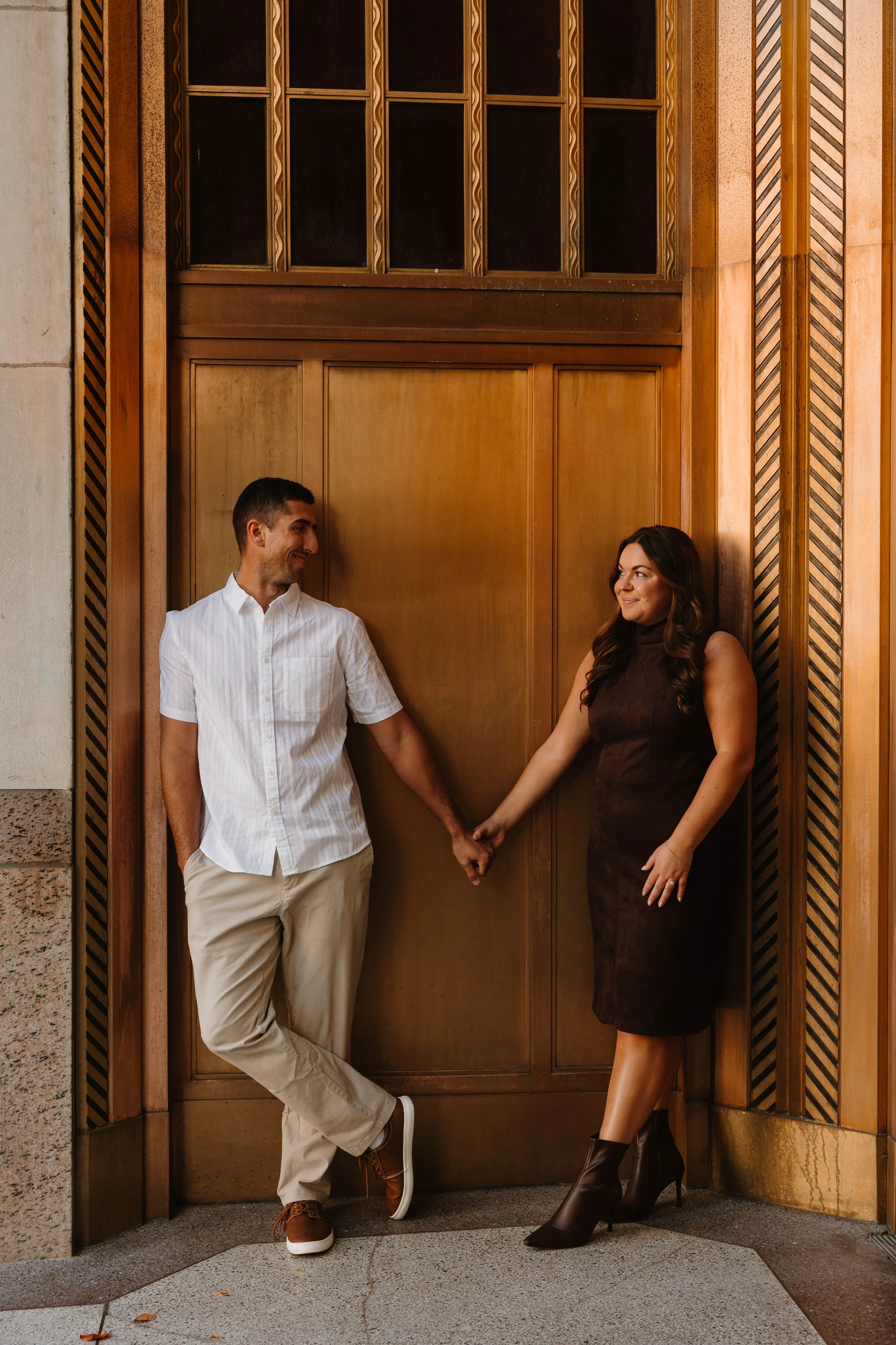 A man and woman hold hands while standing against a large wooden door, looking at each other inside a building with wood-paneled walls.