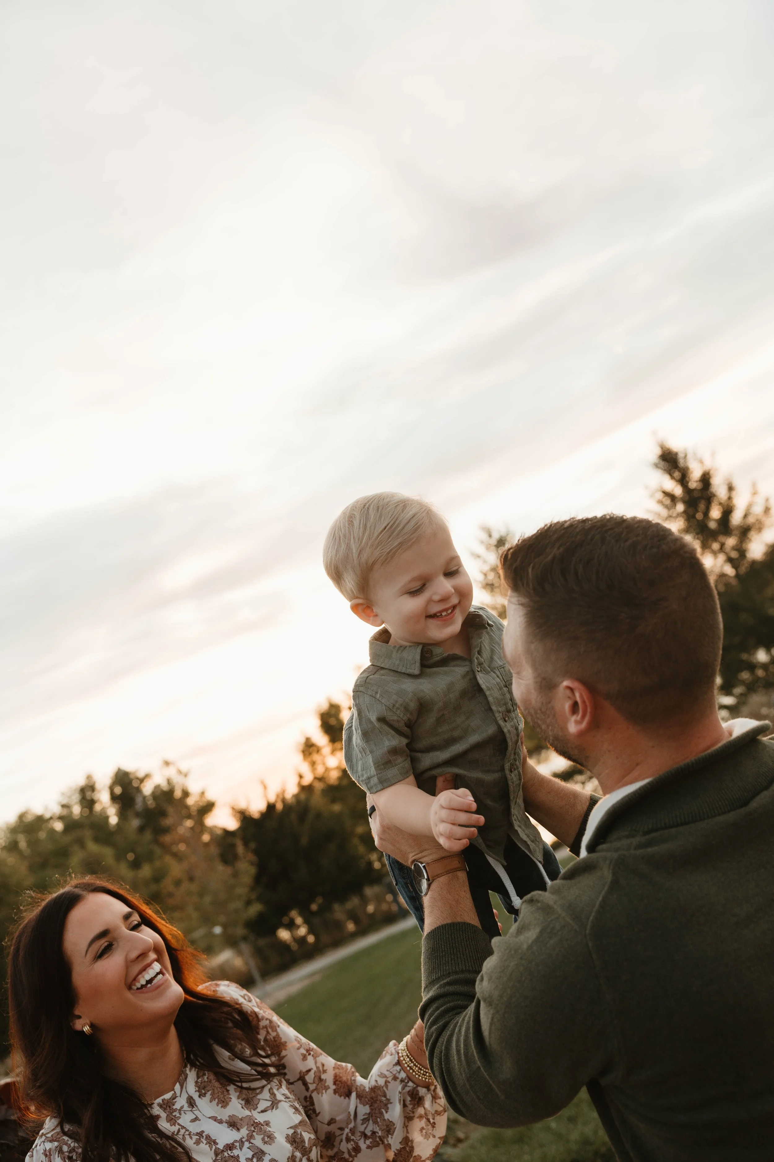 A happy family outdoors during sunset, with a woman smiling, a man lifting a young boy into the air, trees in the background.