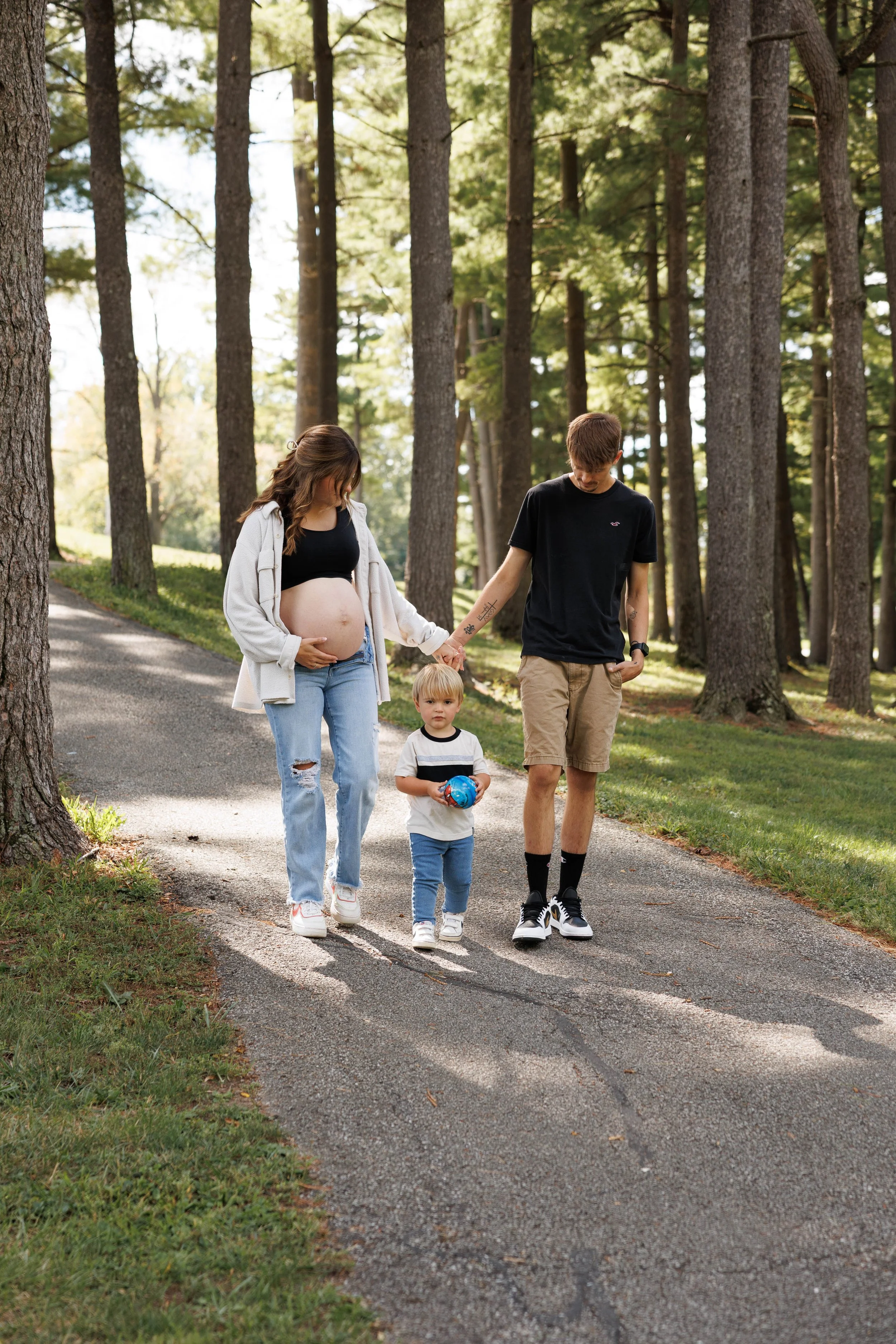 A pregnant woman, a man, and a young child walk together along a paved trail through a wooded park on a sunny day.