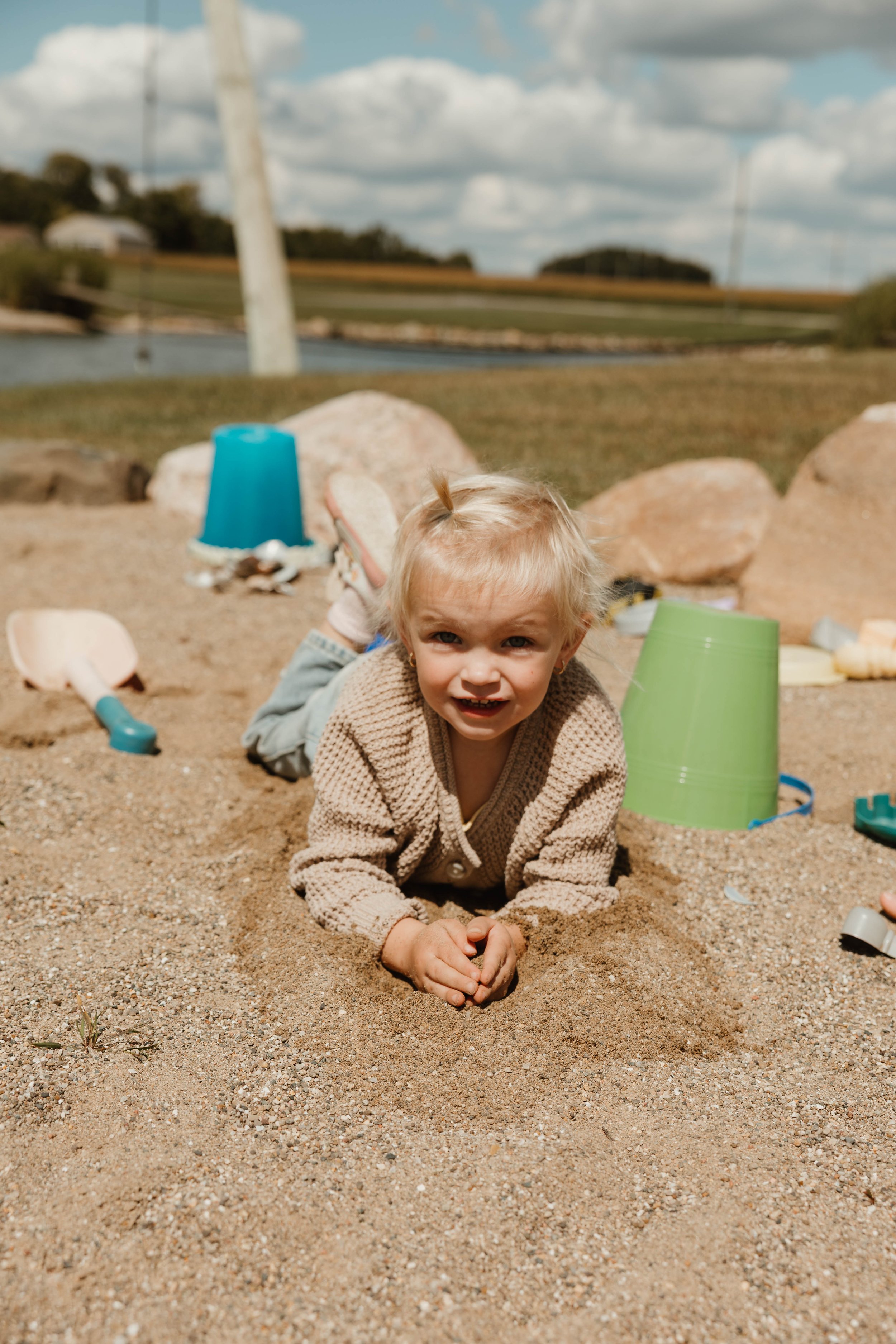A young child playing in the sand at a beach, lying on their stomach with toys around, cloudy sky in the background.
