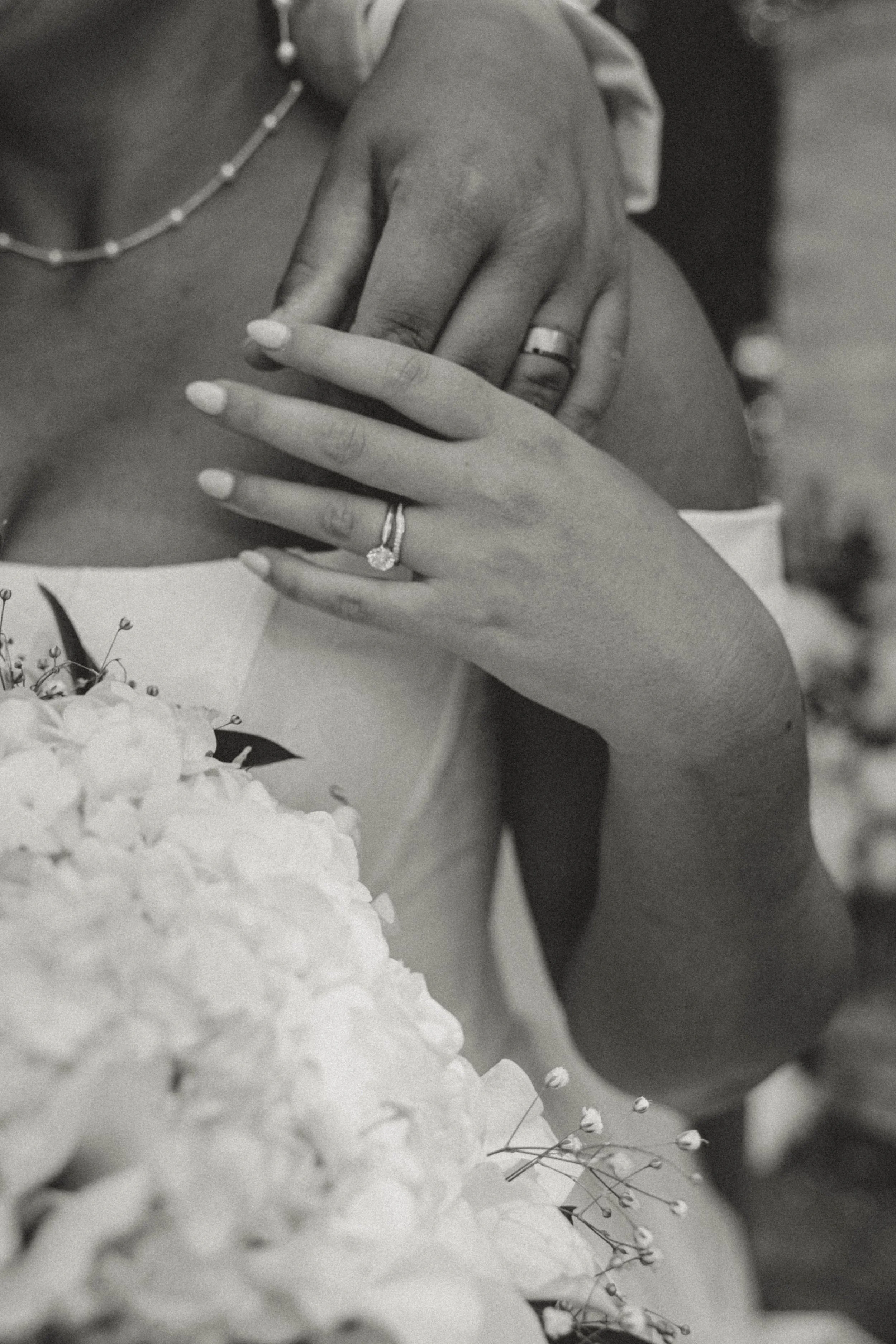 Close-up of a bride and groom's wedding rings on their hands resting on each other's shoulders, with a wedding bouquet in the foreground and the bride wearing a pearl necklace.
