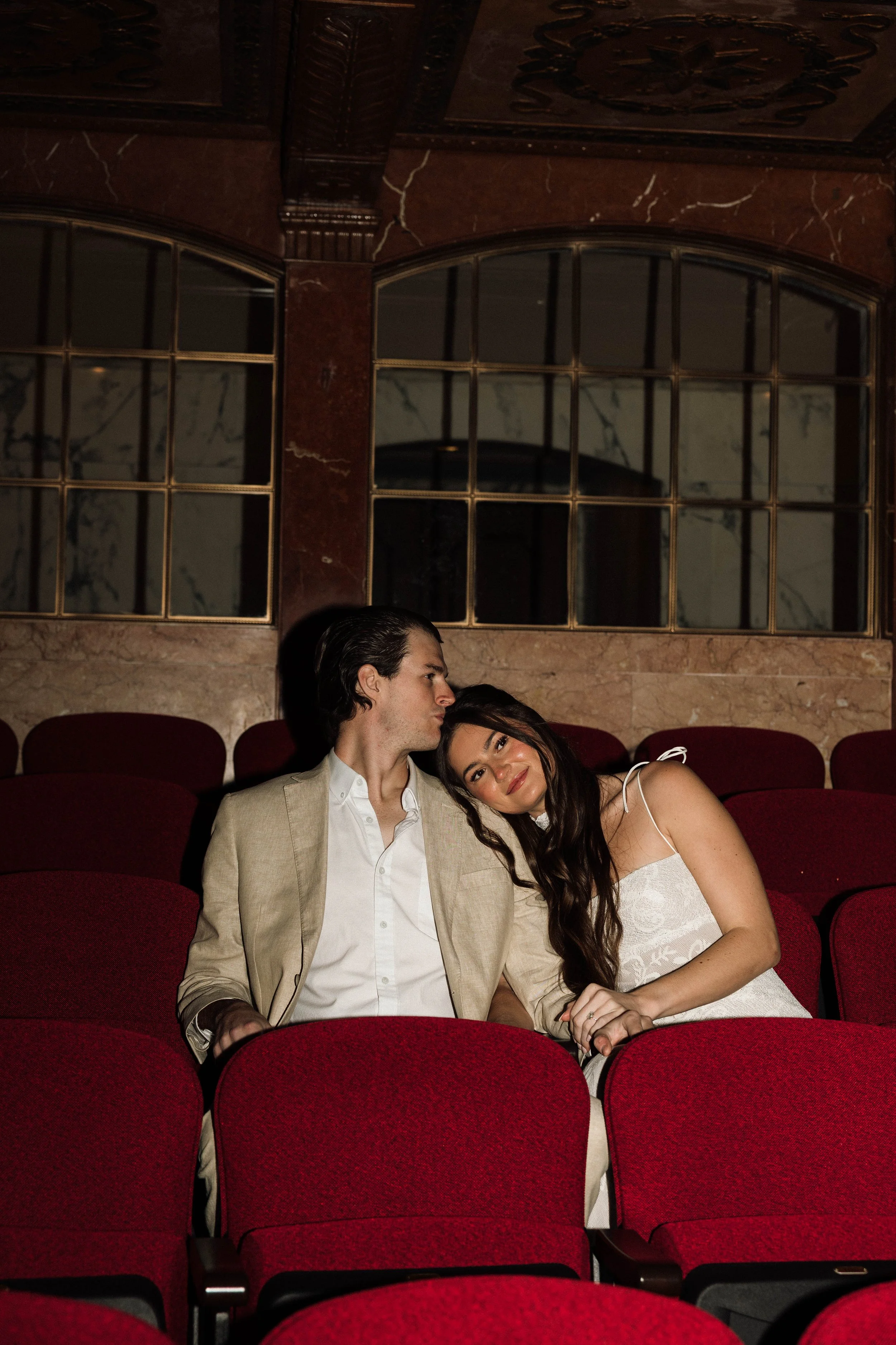 A couple sitting closely together in red theatre chairs inside an ornate, marble-walled auditorium. The woman is leaning her head on the man's shoulder, smiling gently, while the man looks at her with a soft expression.