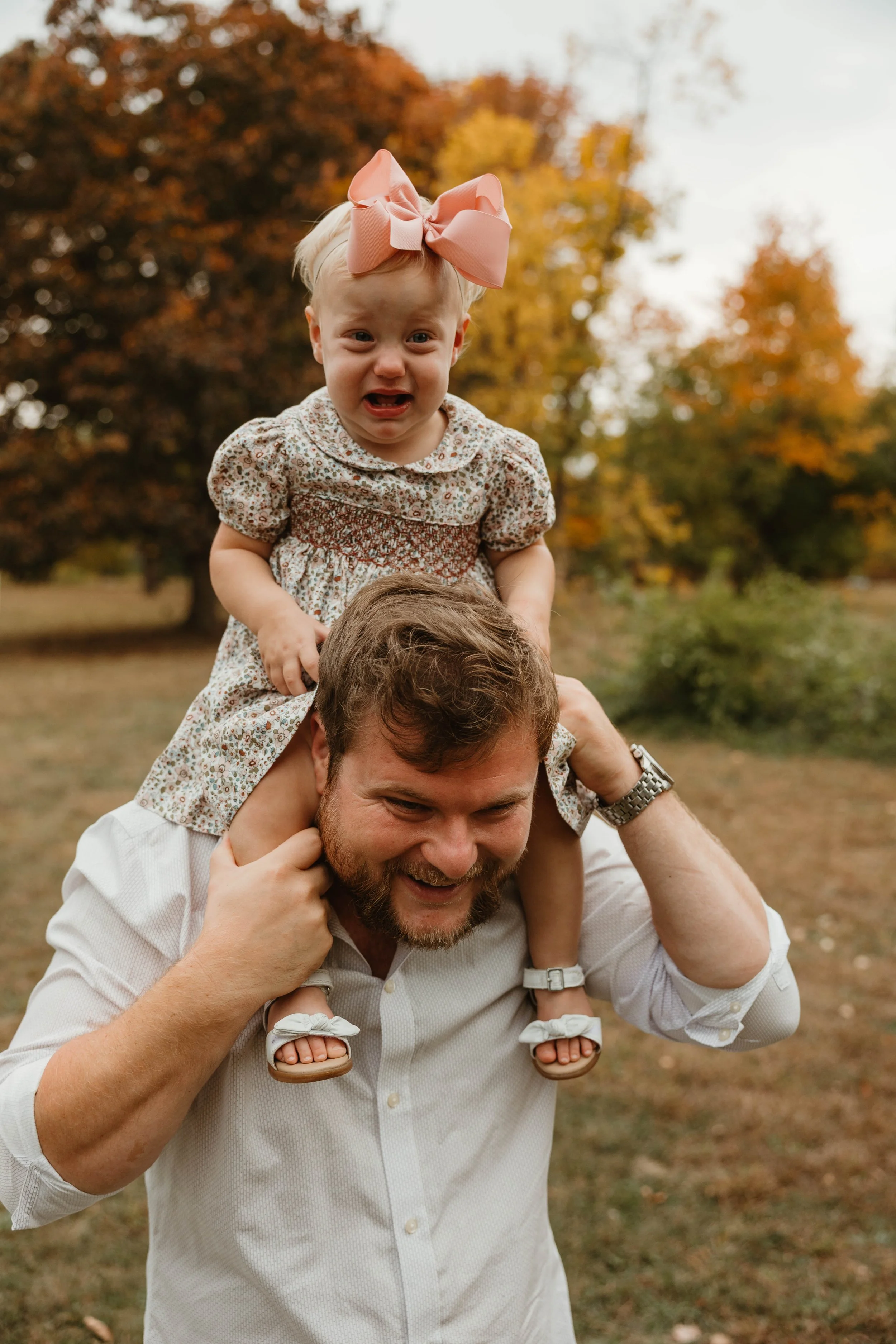 A man with a beard and white shirt is smiling as he carries a young girl on his shoulders. The girl has blonde hair, a pink bow, and is wearing a floral dress. They are outdoors in a park with fall foliage in the background.