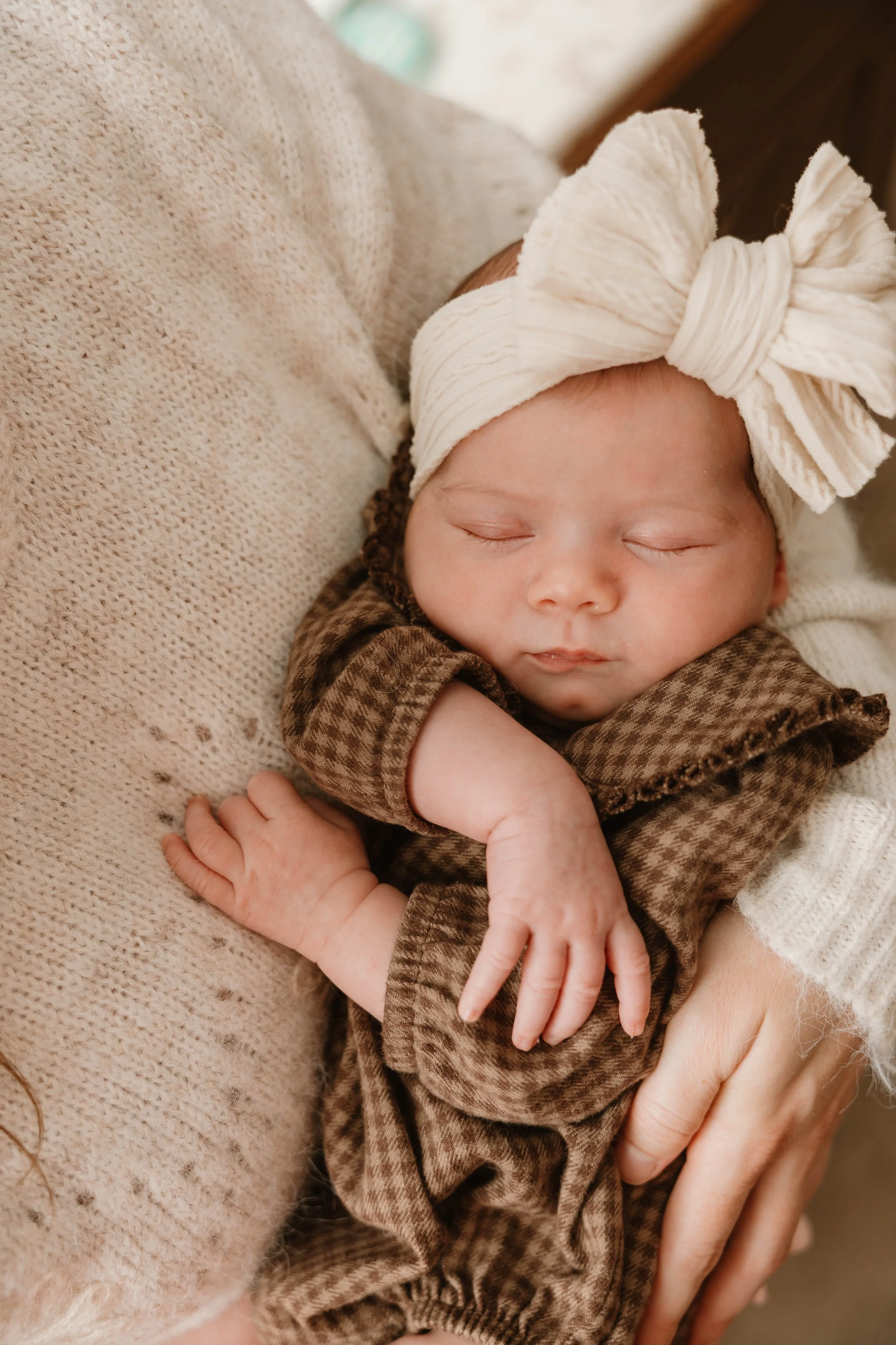 A sleeping baby girl wearing a cream headwrap with a bow and a brown checkered outfit, resting on an adult's arm, on a beige knitted blanket.