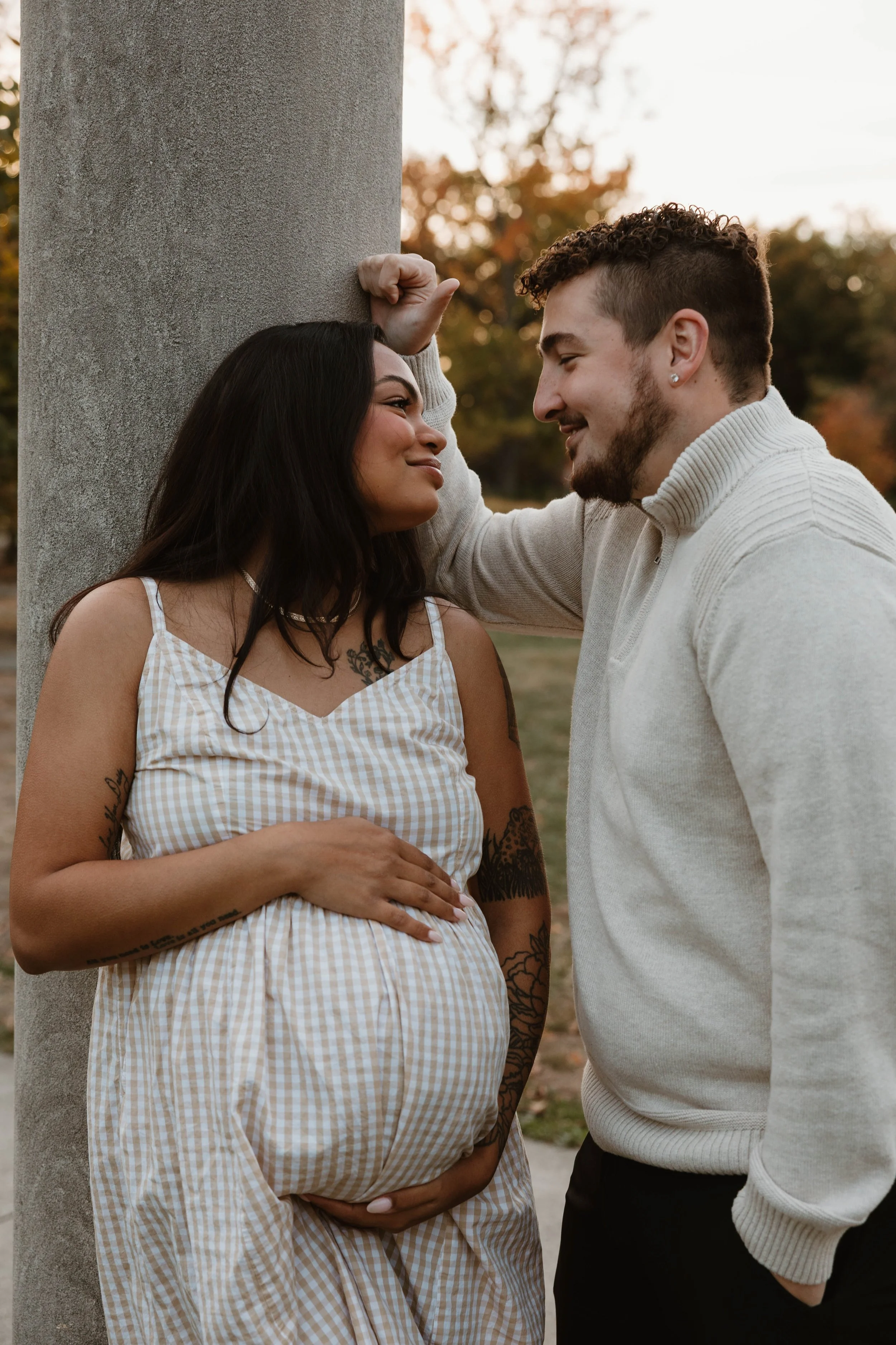 A pregnant woman and a man standing close, smiling at each other, leaning against a concrete pillar outdoors during fall.