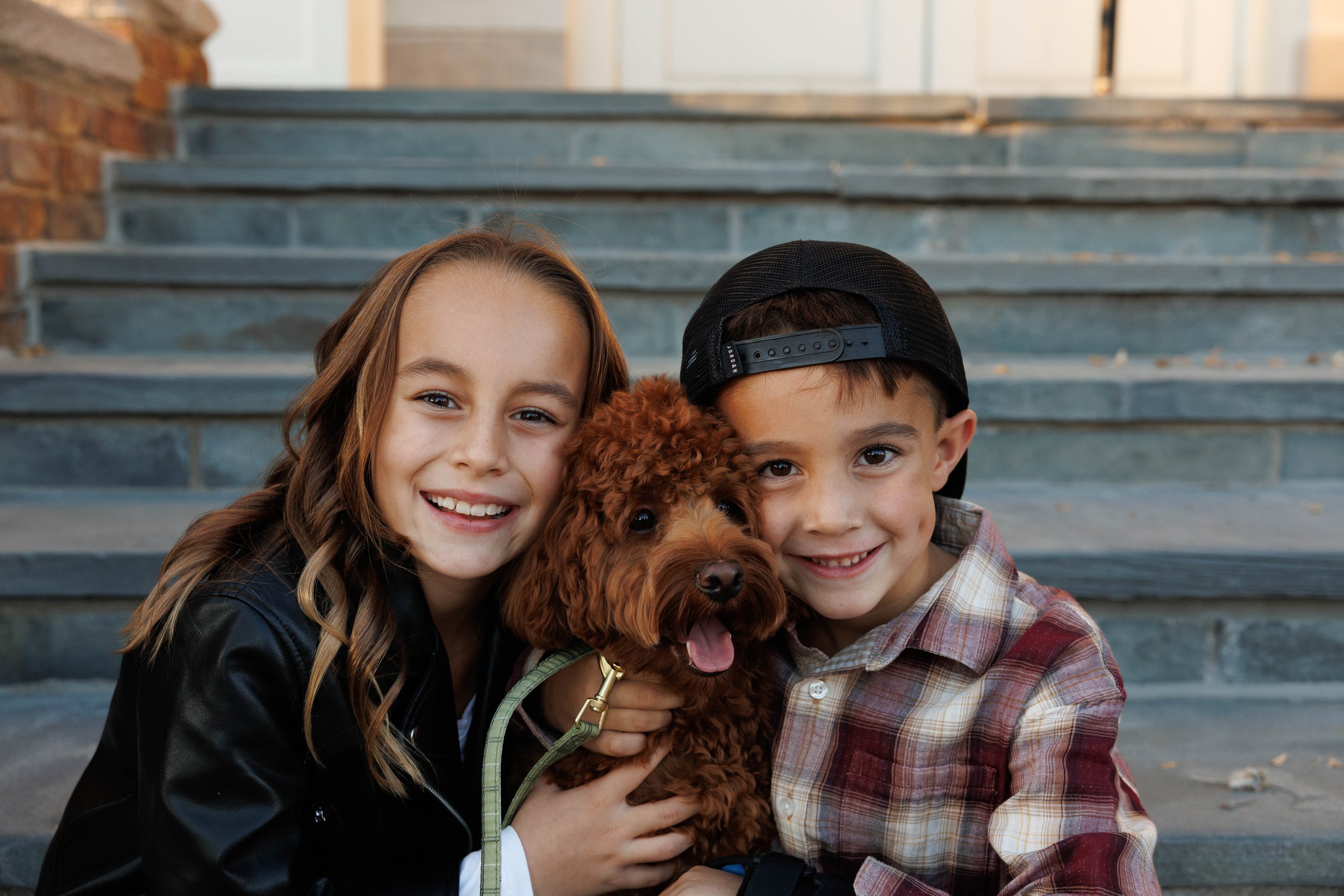 Two kids, a girl and a boy, smiling and holding a brown toy poodle puppy, sitting on gray stone stairs outside.