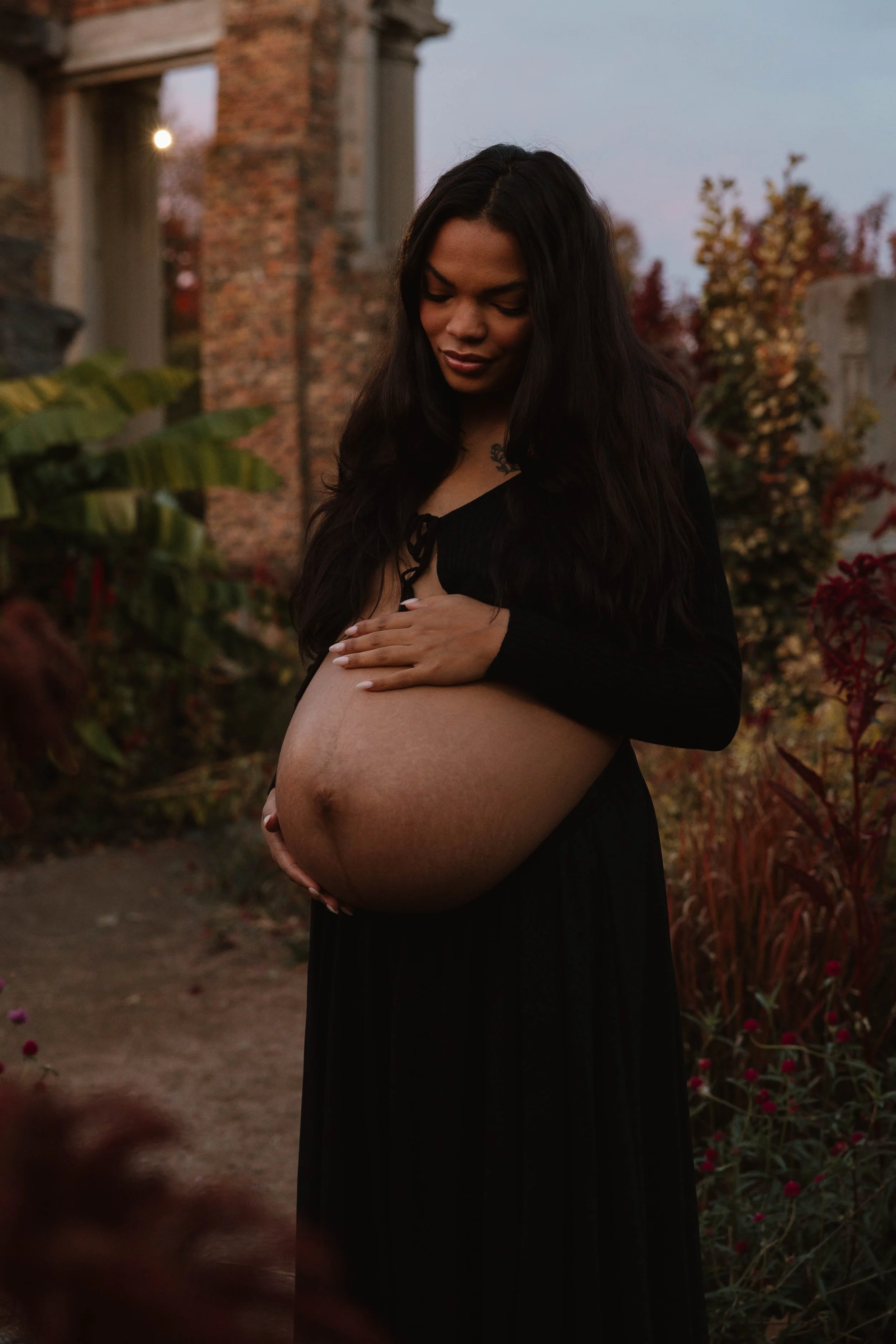 A pregnant woman with long dark hair standing outdoors during sunset, gently holding her belly, surrounded by autumn-colored plants and ruins in the background.