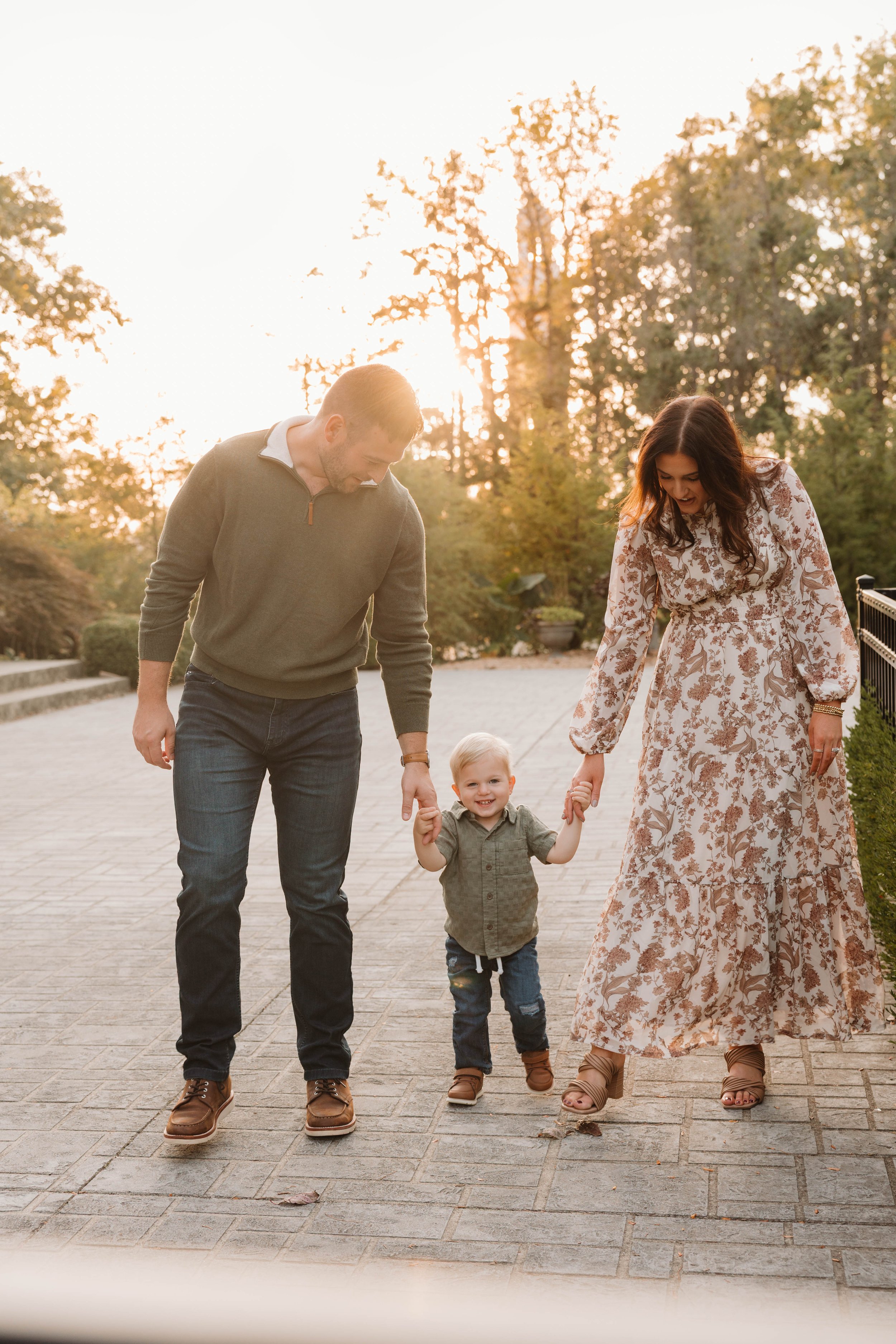 A family of three, a man, a woman, and a young boy, walking outdoors on a sunny day. The man and woman are holding the boy's hands, and they are smiling. The background shows trees and a paved pathway.