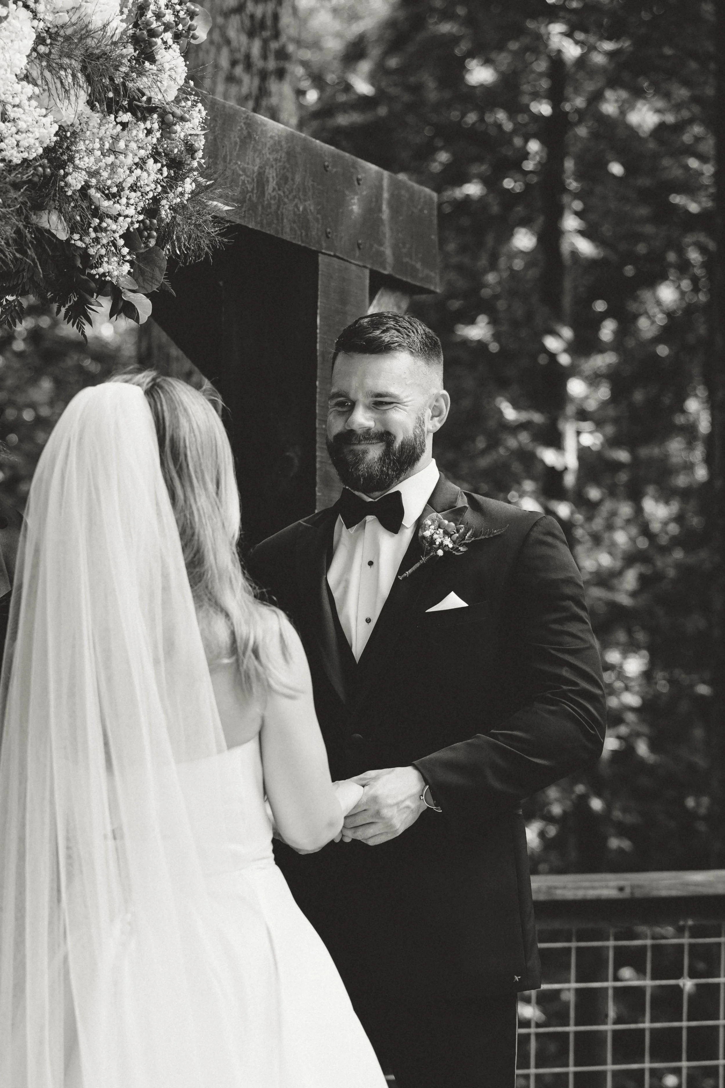 A black and white photo of a bride and groom during their outdoor wedding ceremony. The groom is smiling and wearing a tuxedo with a bow tie, while the bride, in a white wedding dress and veil, faces him, holding hands.