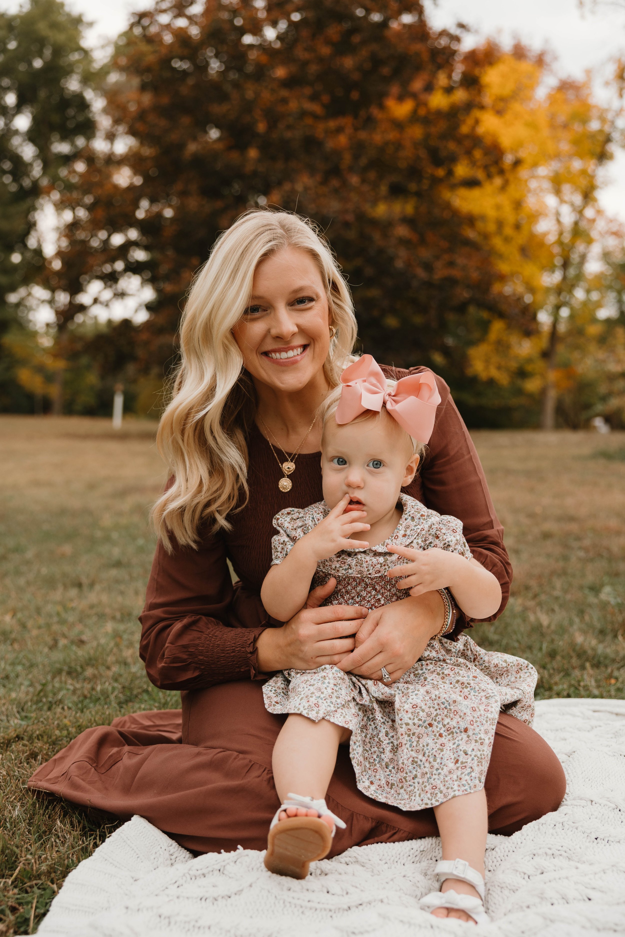 A woman with blonde hair and a little girl with blonde hair, both sitting outdoors on a white blanket during autumn. The woman is smiling and holding the little girl on her lap, who has a pink bow in her hair and is touching her mouth with her finger