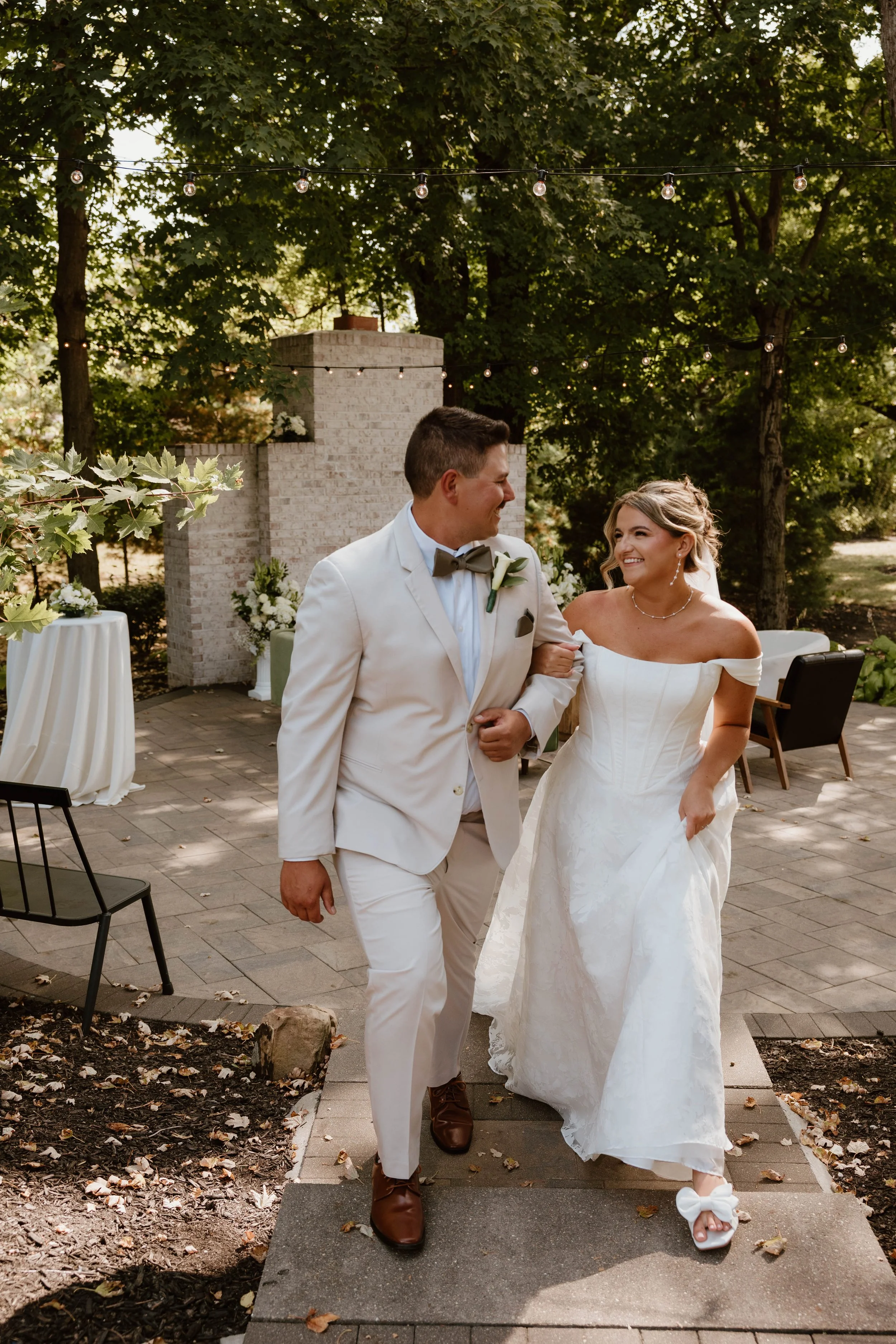 A bride and groom walking arm in arm outdoors at a wedding, smiling at each other, under string lights, with trees and wedding decor in the background.
