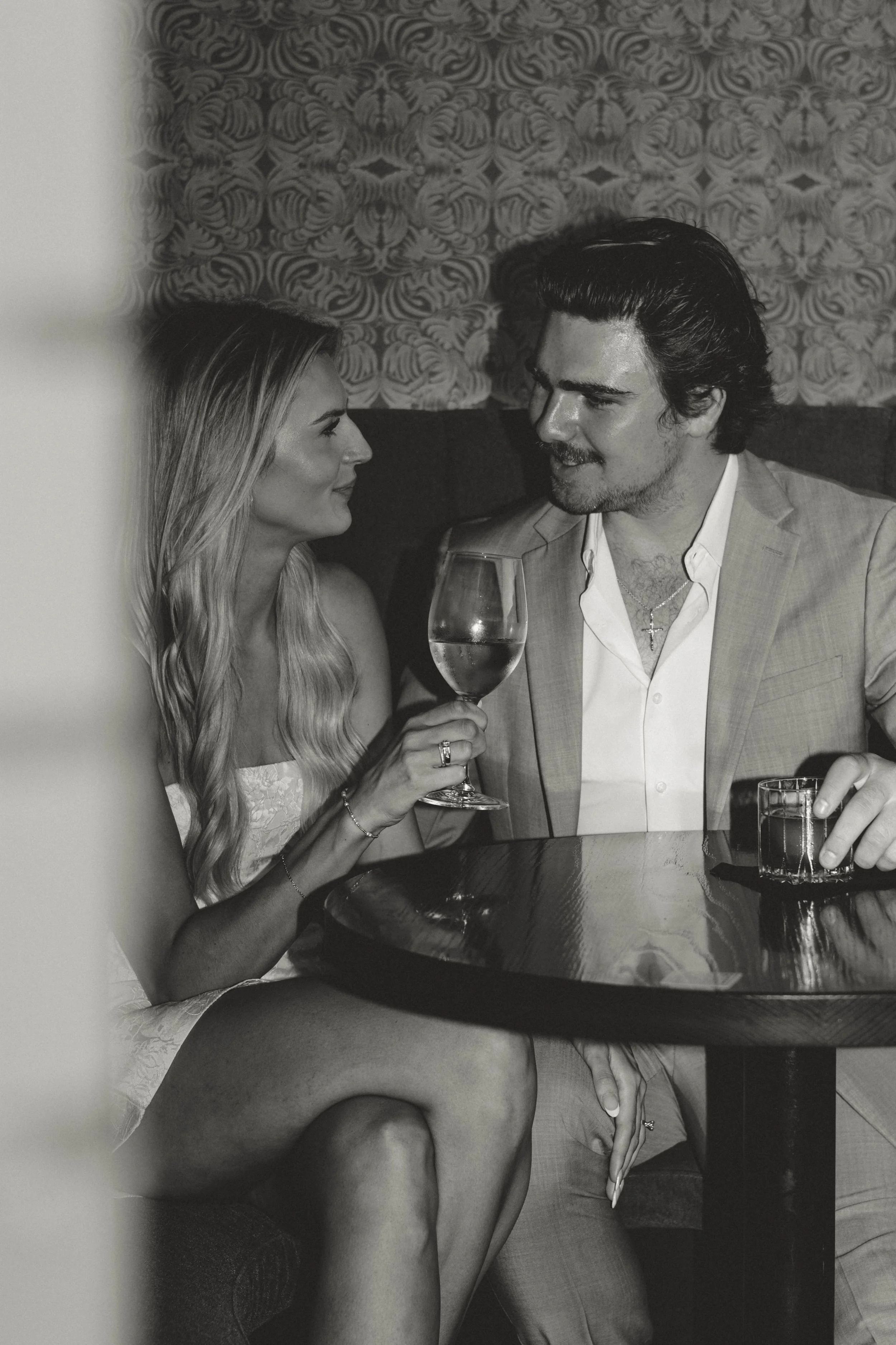 A man and woman sitting at a bar or restaurant table, engaging in conversation. The woman is holding a glass of wine, and the man has a glass of whiskey on the table. They are looking at each other, smiling, with a patterned wallpaper background.