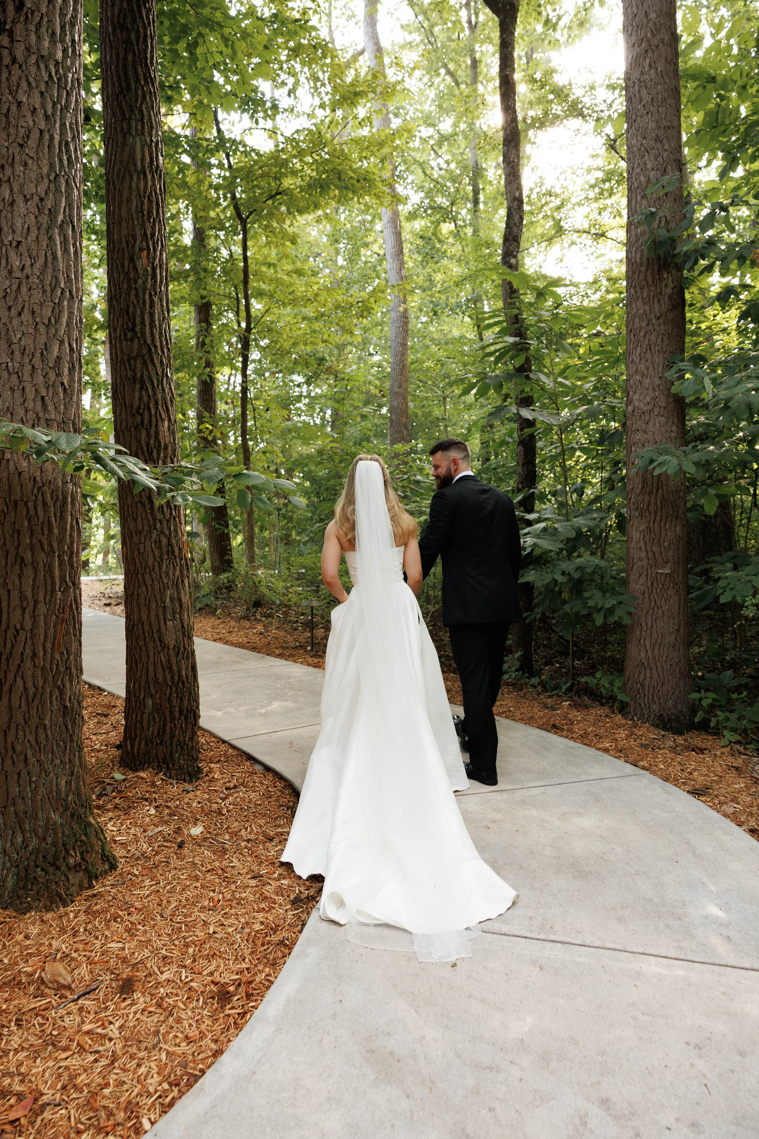 A bride and groom walking hand in hand on a forested path, with tall trees and green foliage surrounding them in a natural outdoor setting.