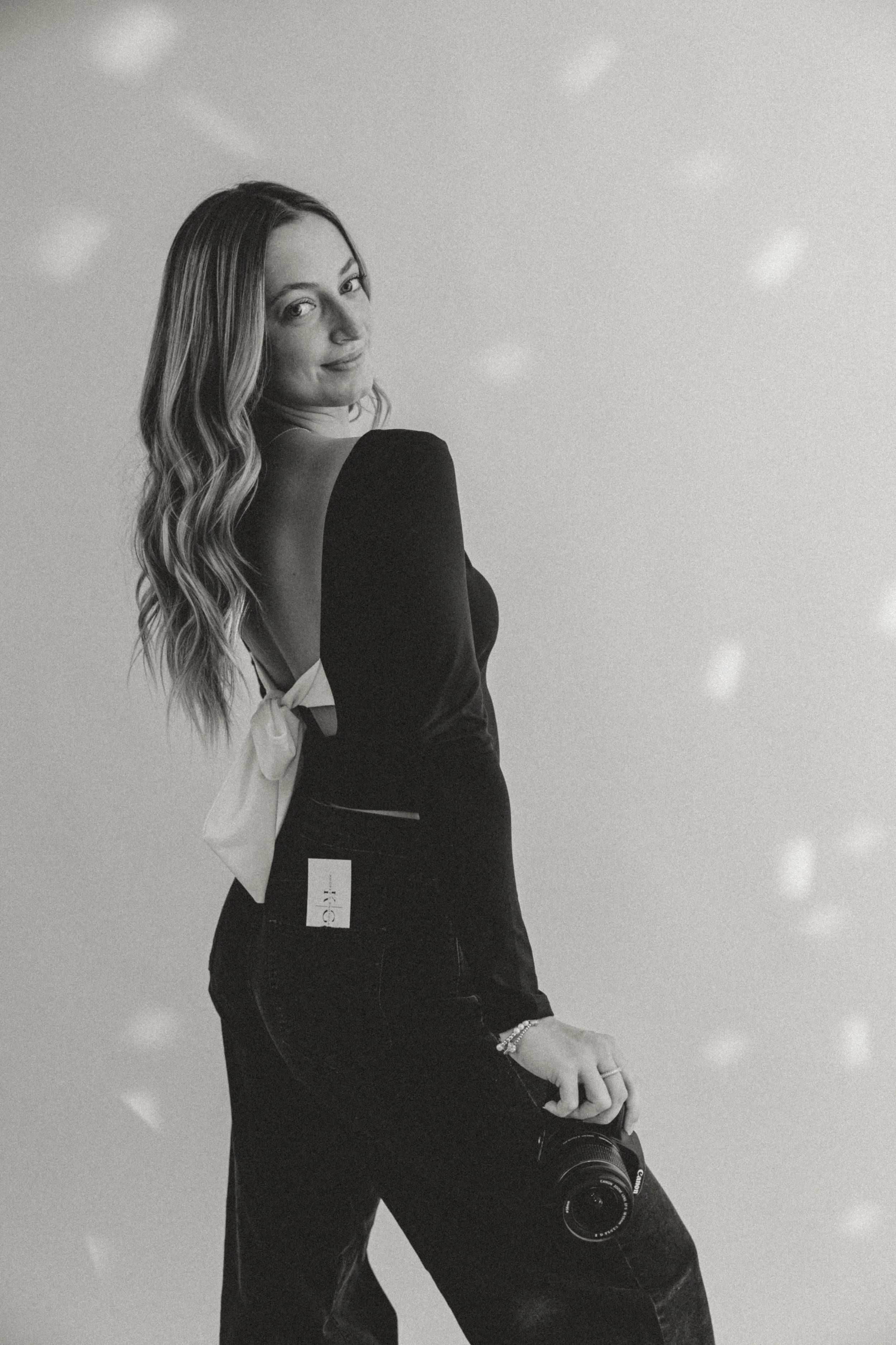 A woman with wavy hair looking over her shoulder, taking a selfie in front of a plain background with light spots, holding a camera in one hand.