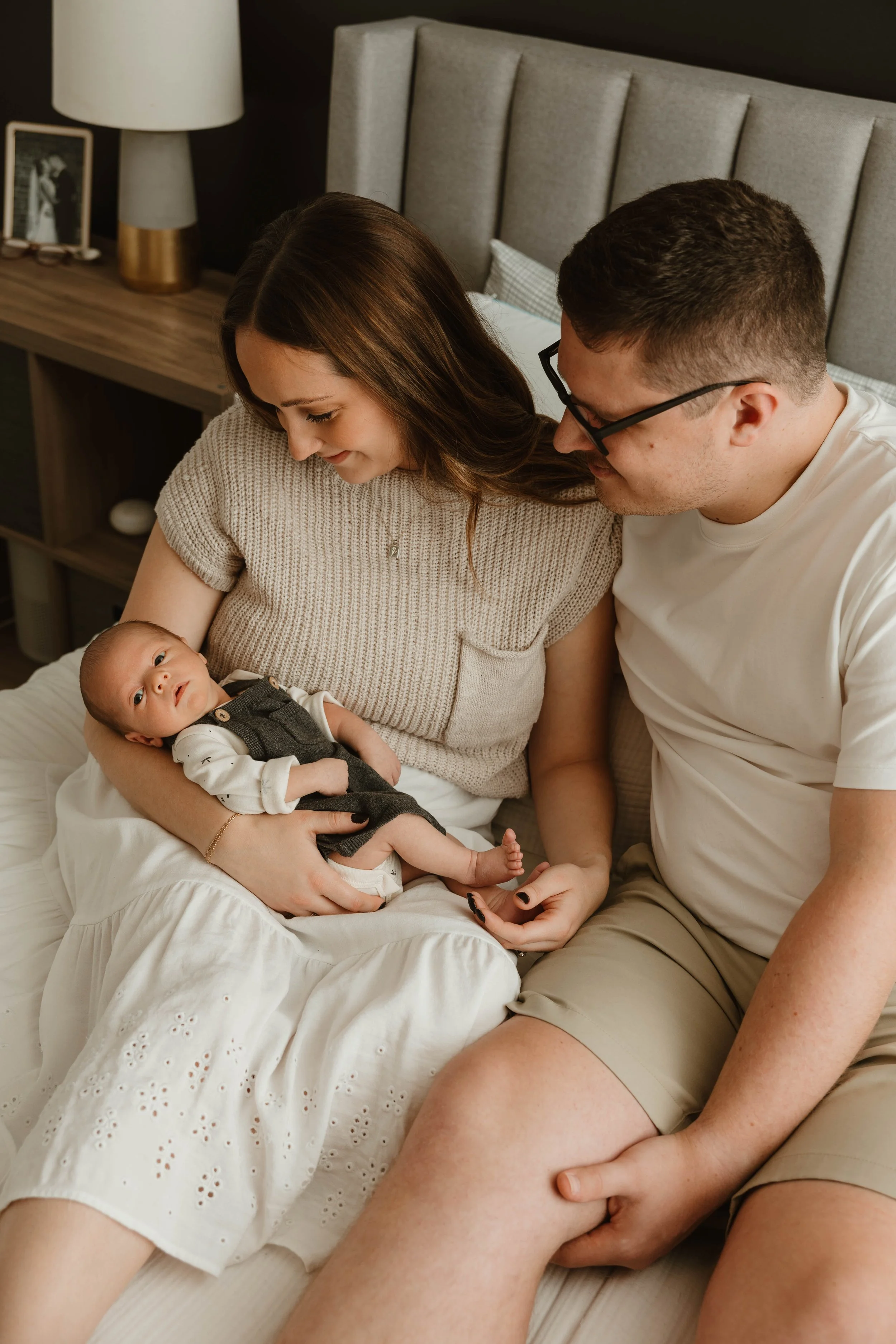 A family sitting on a bed with a woman holding a newborn baby, a man sitting close to her, in a cozy bedroom.