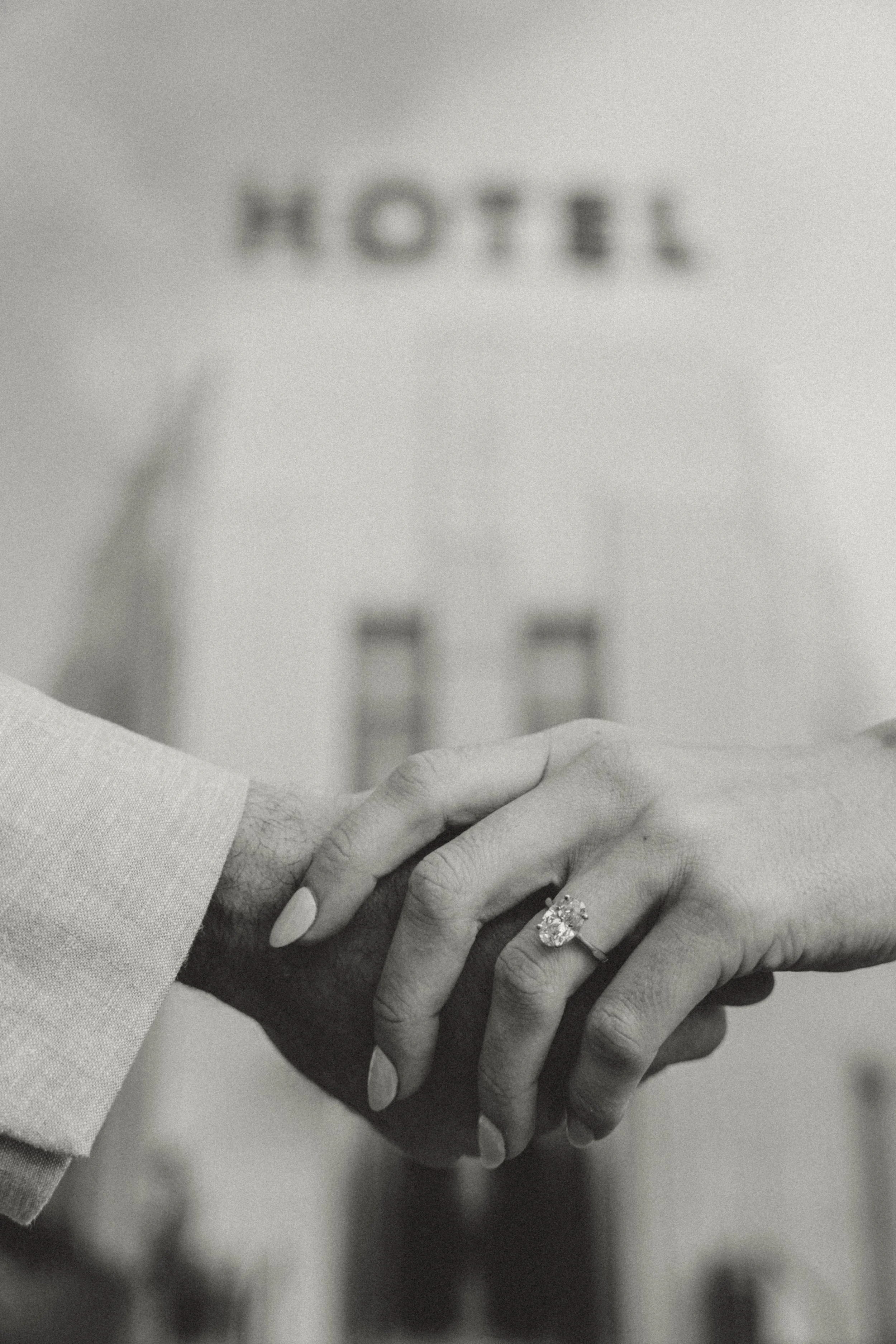 Close-up of hands holding with a wedding ring on the woman's finger, with a blurred sign reading 'HOTEL' in the background.
