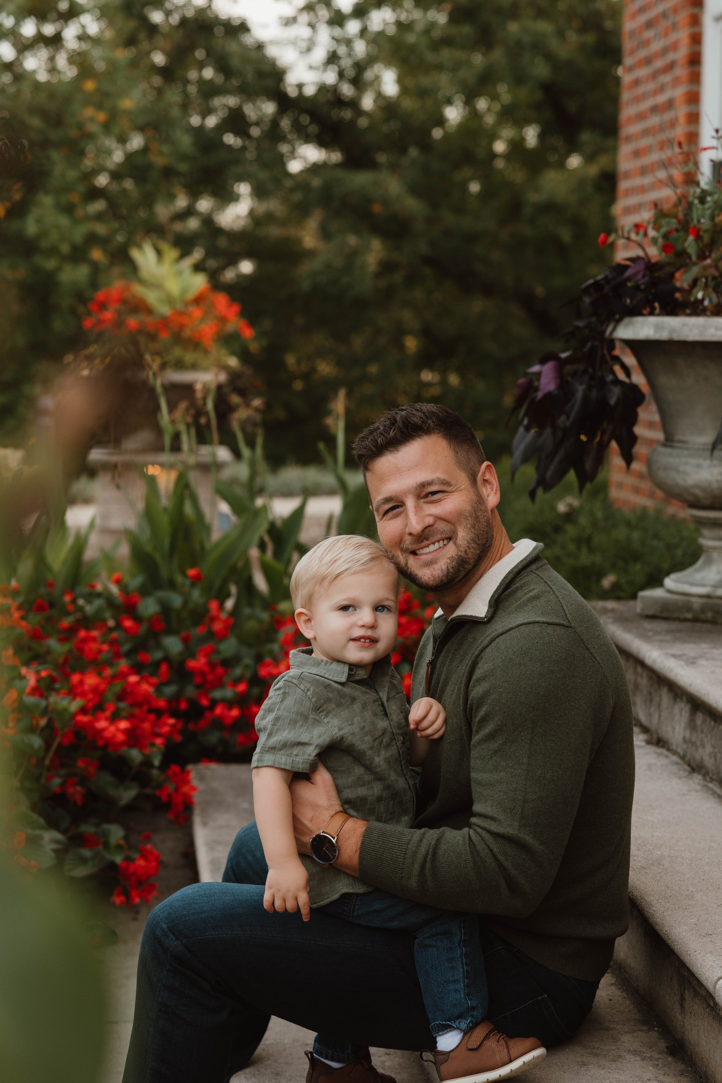 A man and a young boy sitting on outdoor steps with greenery and flowers around them, smiling at the camera.