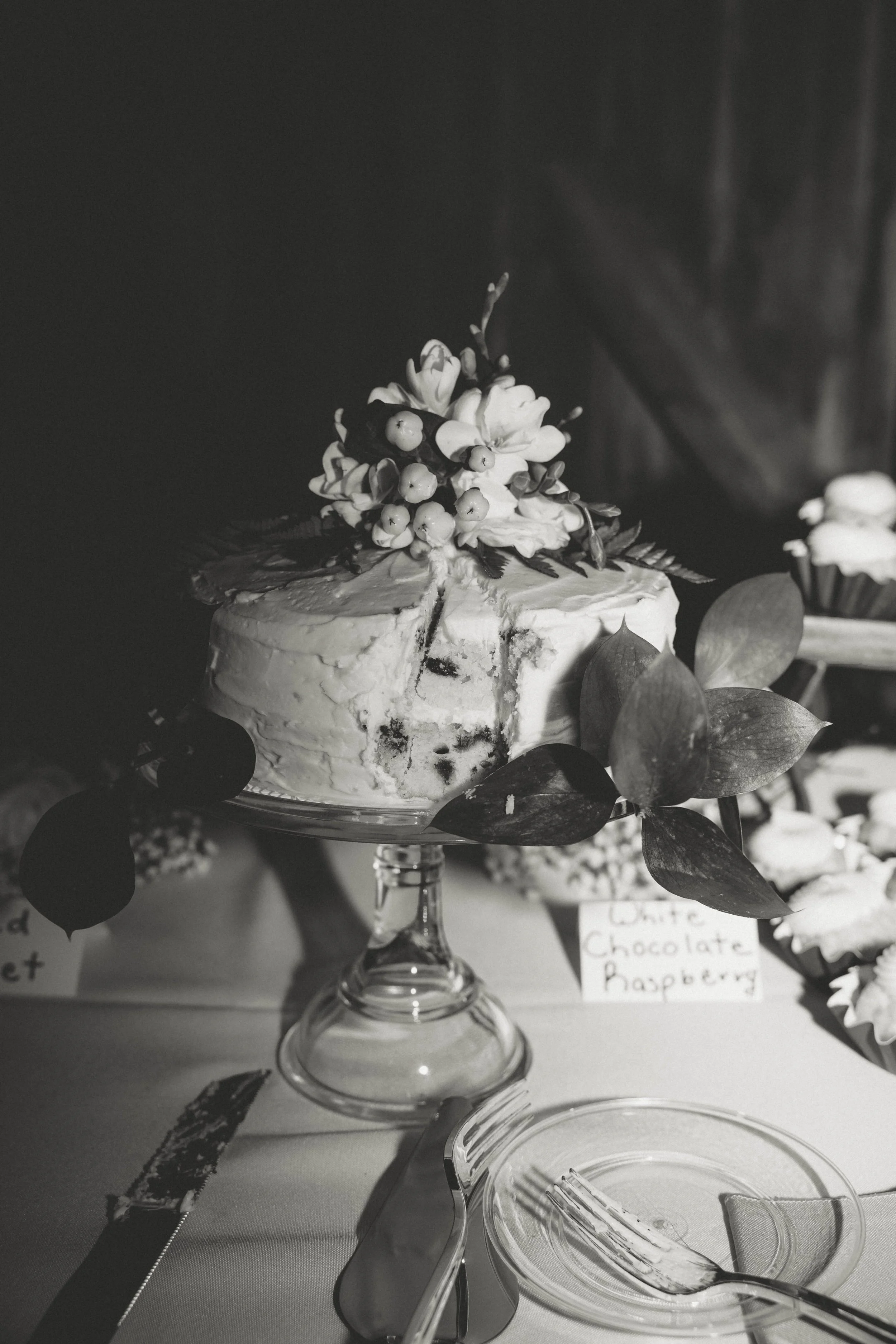 A partially sliced cake topped with flowers and berries, displayed on a glass cake stand with leaves decoration, at a celebration table with utensils and a label that reads "White Chocolate Raspberry."