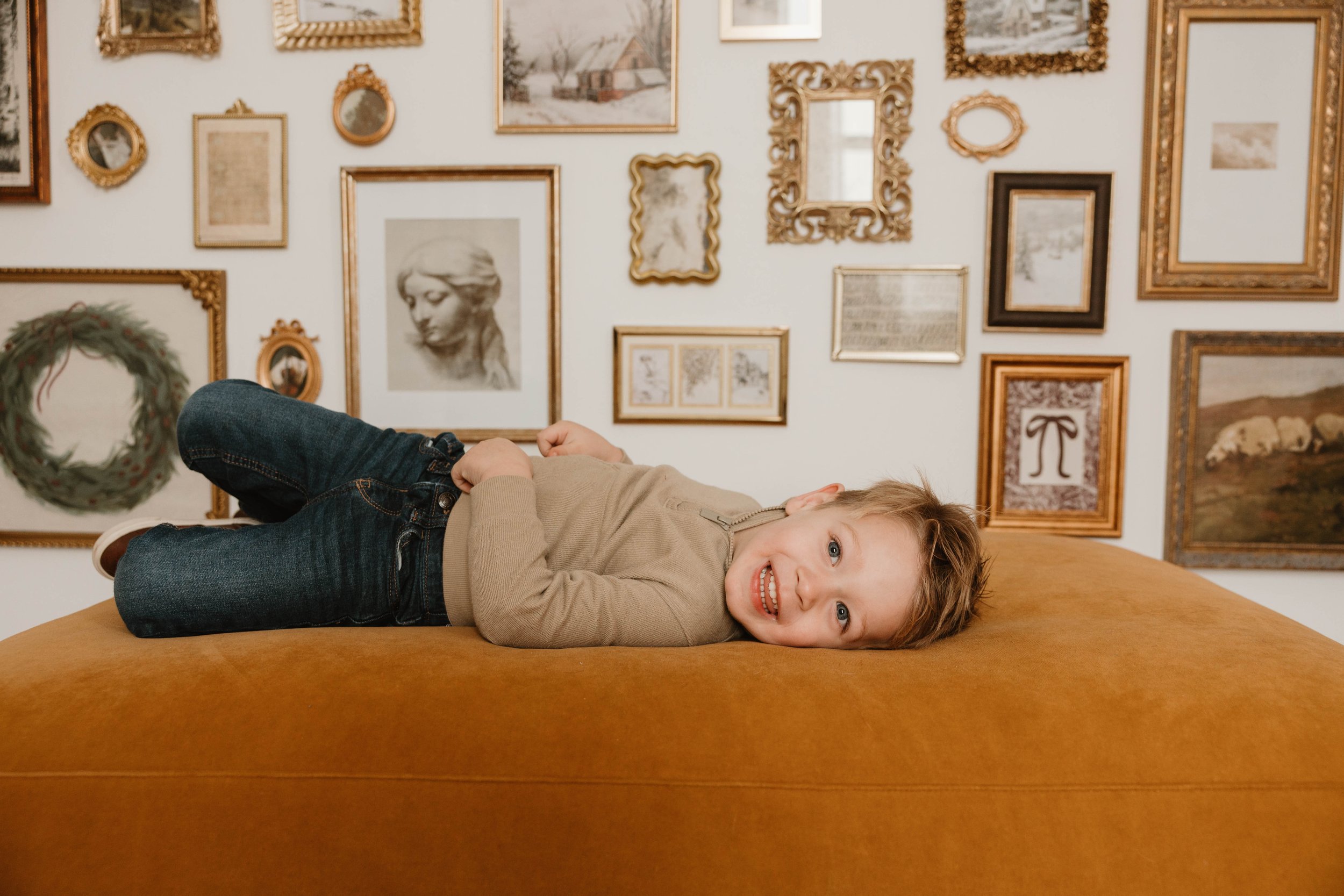 A young boy with light brown hair and blue eyes lying on a mustard-colored ottoman, smiling, with a gallery wall of framed pictures including art, photographs, and mirror behind him.