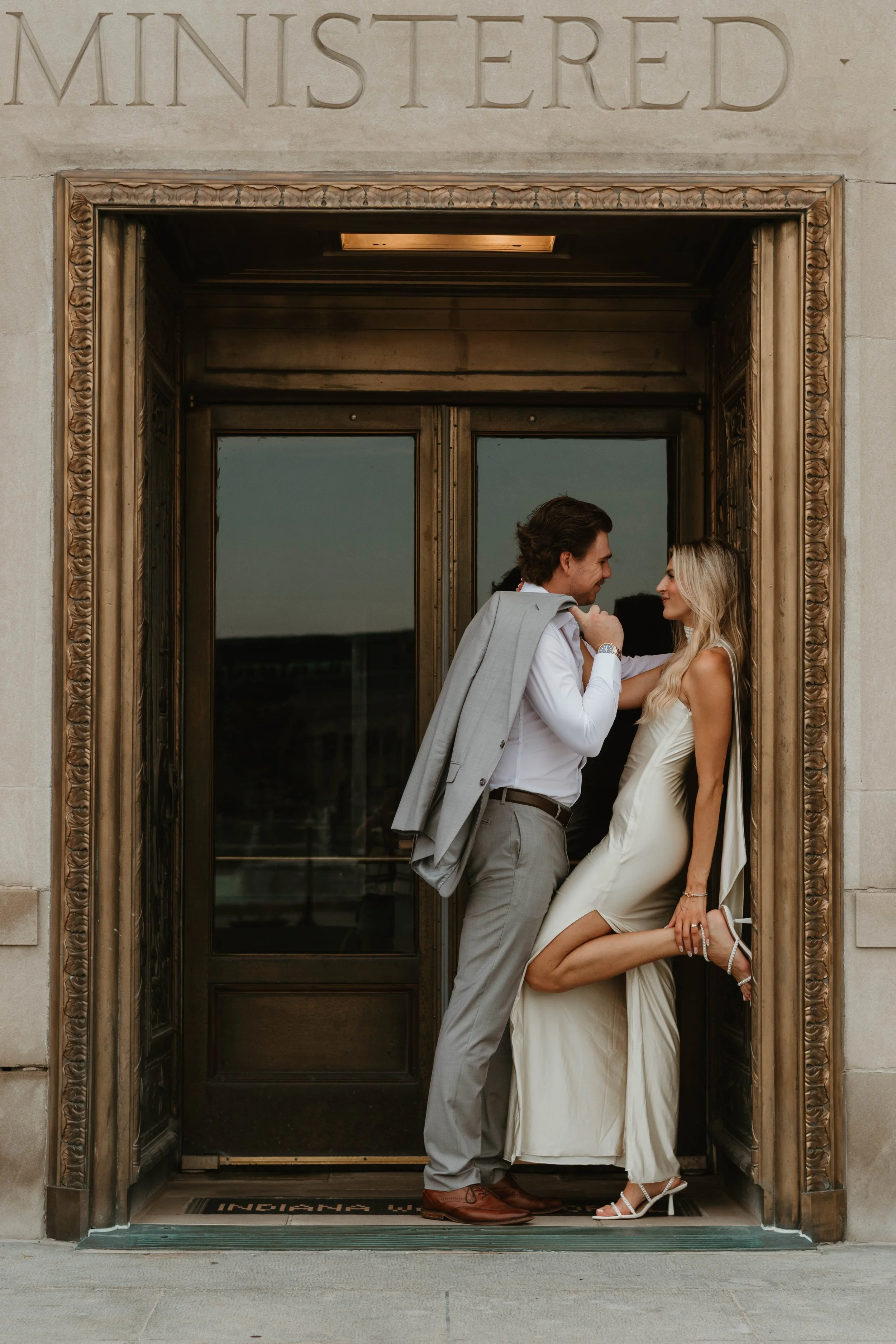 A romantic couple stands close together in a doorway, with the man holding the woman's chin and smiling at her. The woman is wearing a cream-colored dress and high heels, and the man is dressed in a white shirt and gray pants with a jacket draped ove