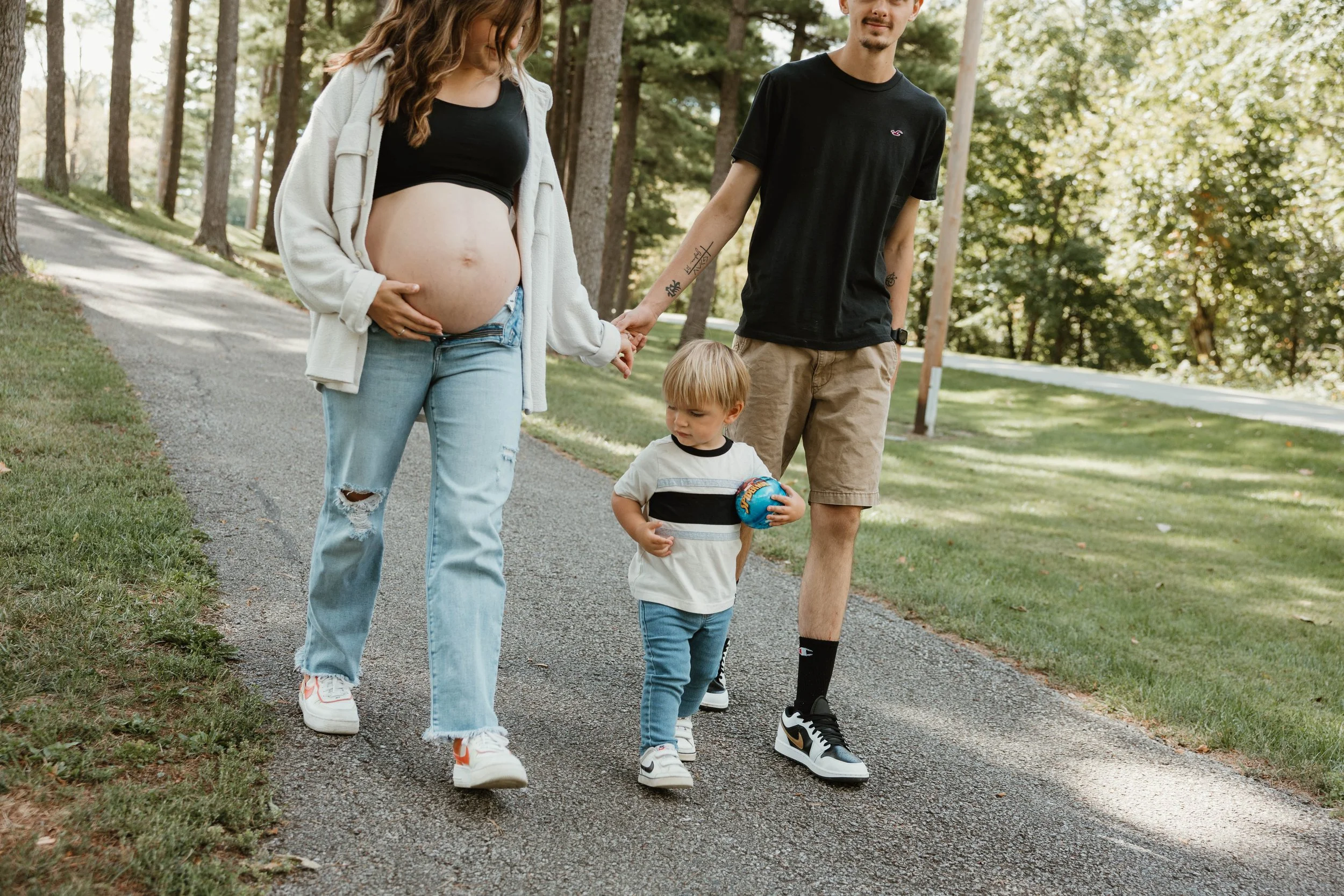 A pregnant woman and a young boy walking on a park path holding hands. The woman is touching her belly. The boy is holding a small blue ball, and a man is walking beside them.