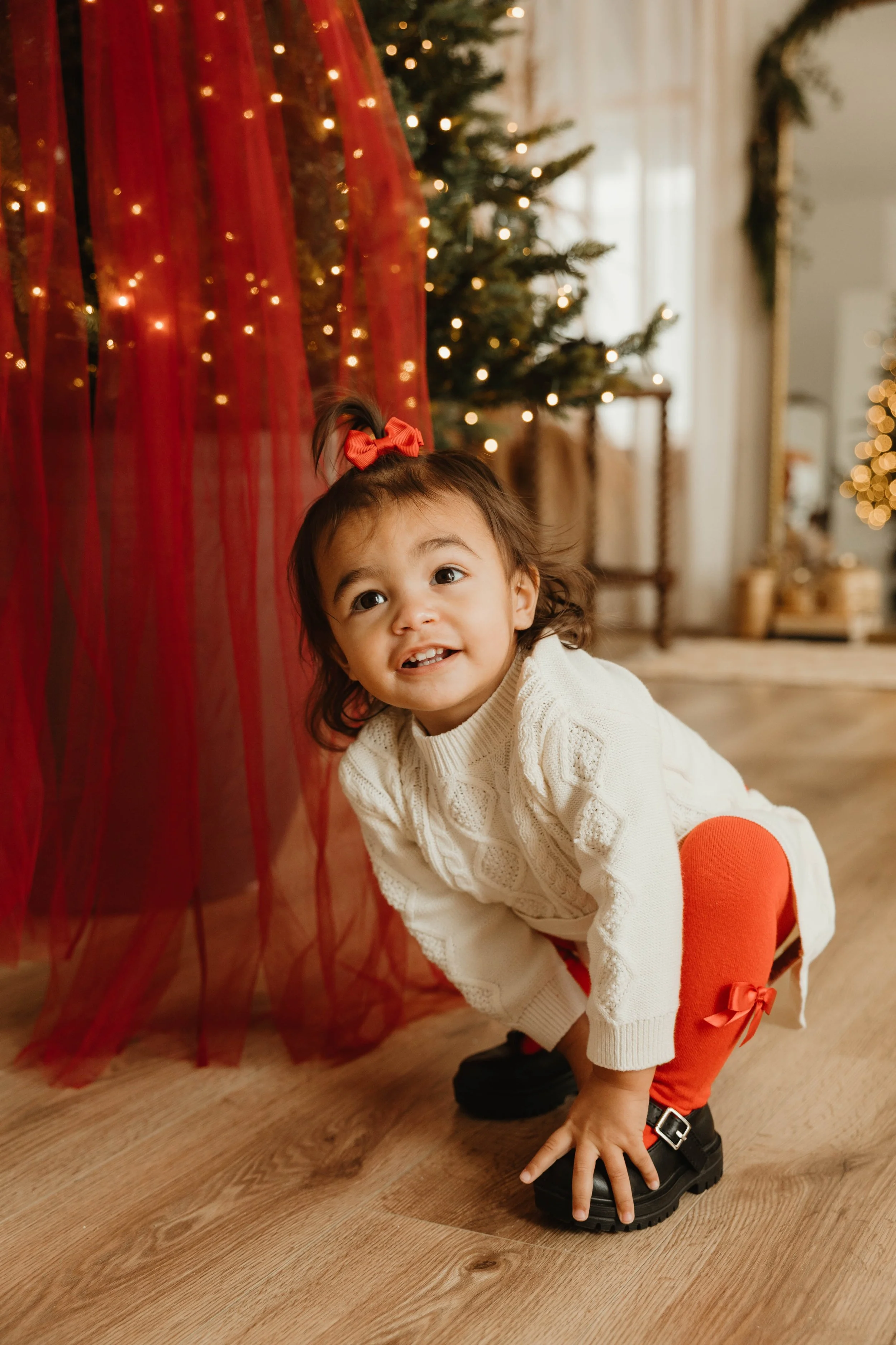 A young girl squatting on a wooden floor, smiling, near a Christmas tree decorated with lights and ornaments, with a red curtain or fabric hanging beside her.