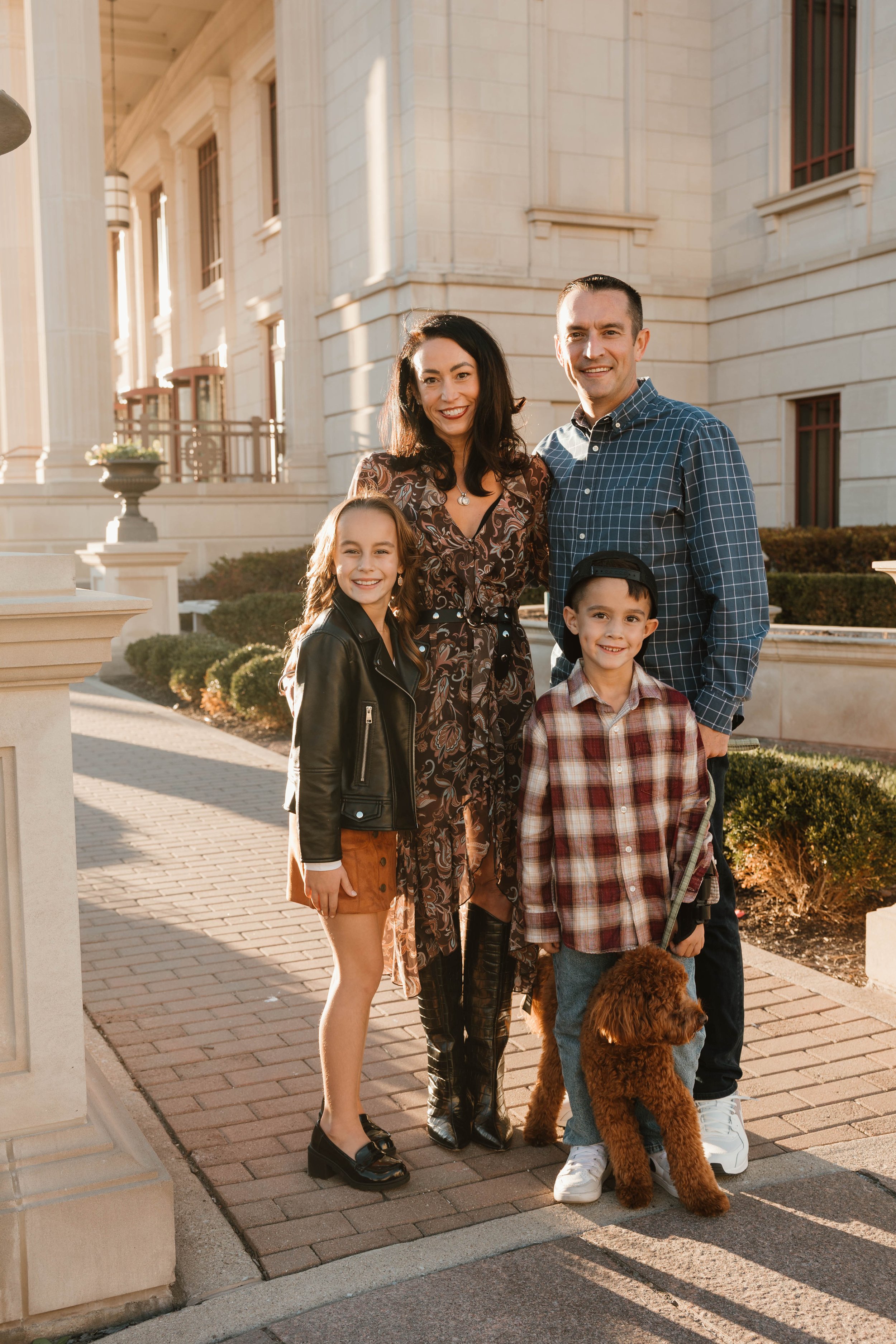 A family of four with their dog standing together outdoors on a sunny day near a large building with classical architecture.
