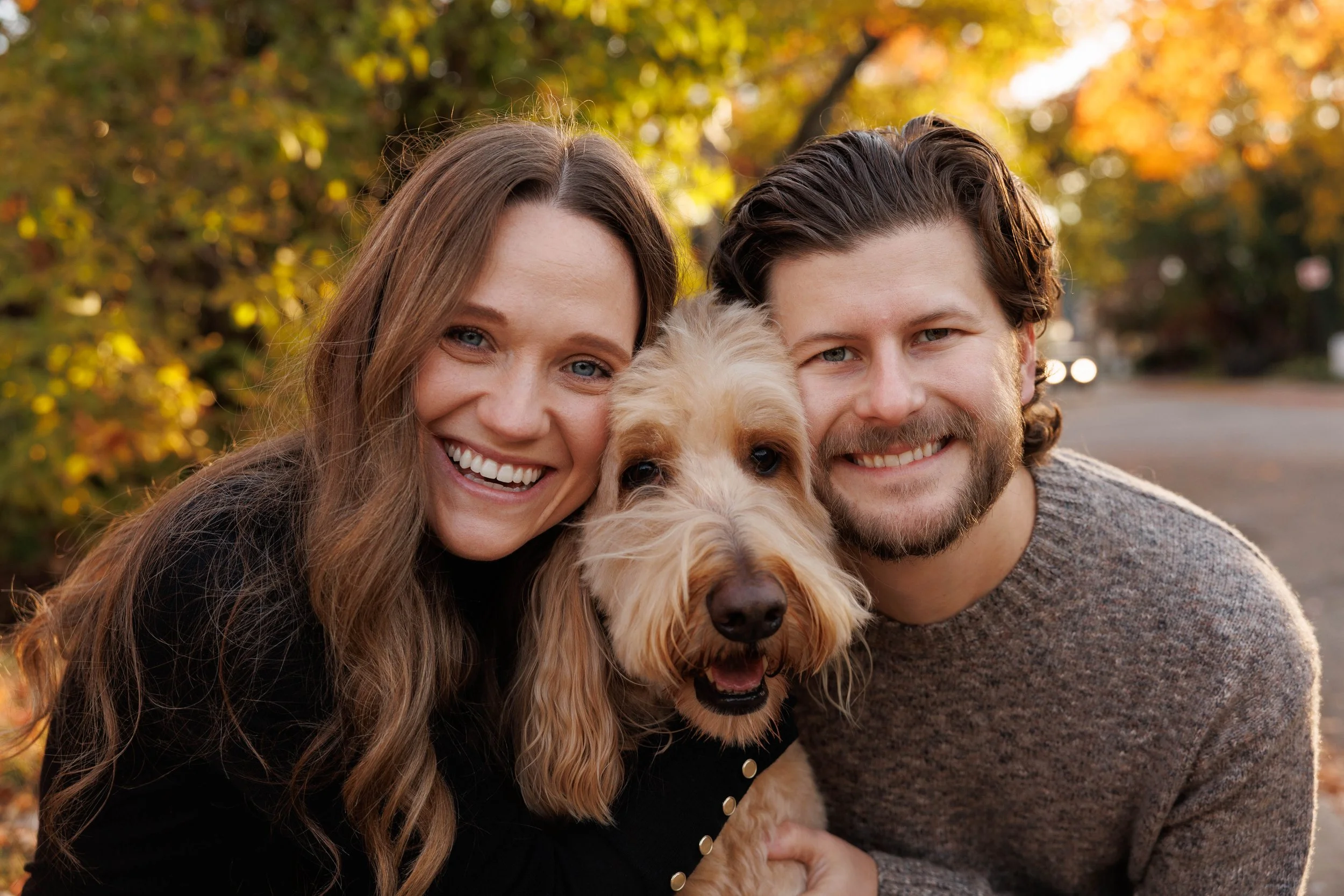 A happy couple with a golden retriever dog outdoors during autumn, surrounded by colorful fall leaves.