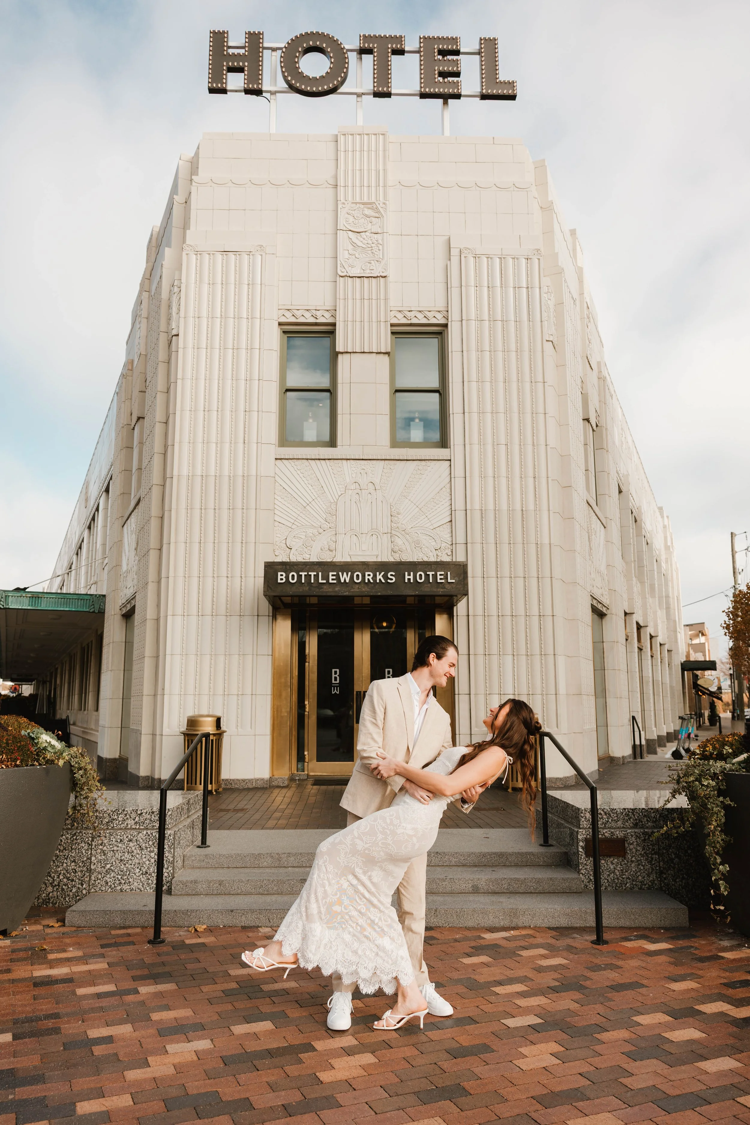 A couple in wedding attire dancing in front of the Bottleworks Hotel, a white Art Deco building, with a large illuminated 'HOTEL' sign on top.