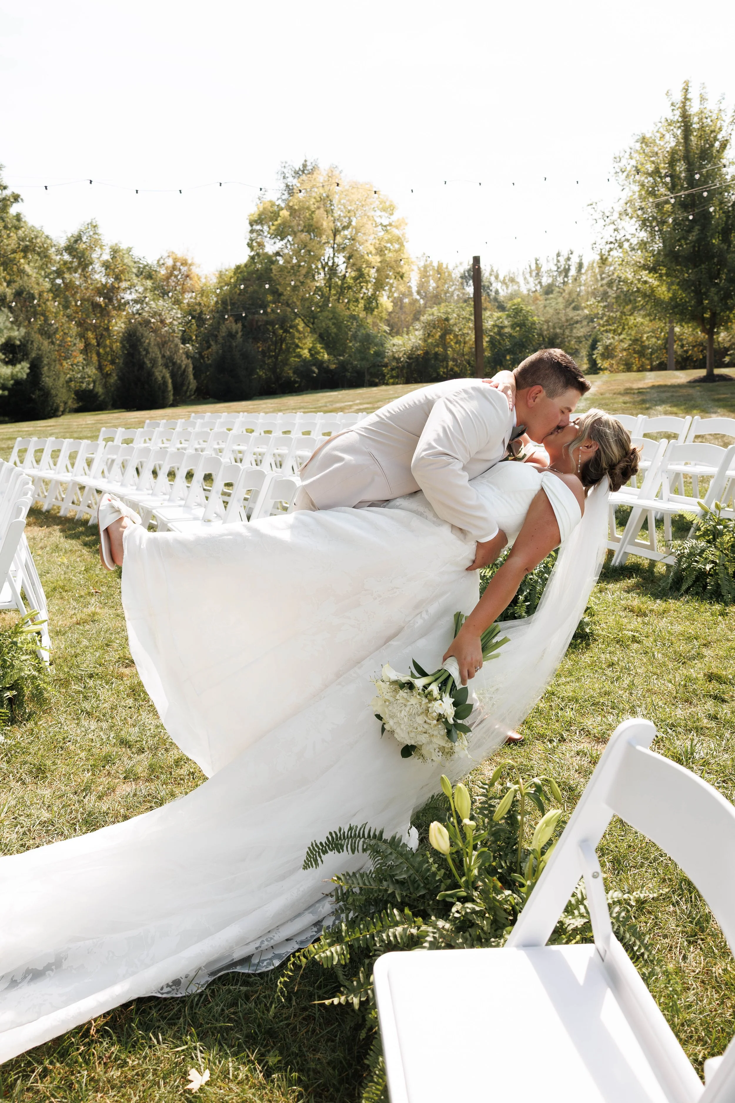 A newlywed couple sharing a kiss outdoors at their wedding ceremony, with the groom lifting the bride in a dip, surrounded by white chairs and greenery on a sunny day.