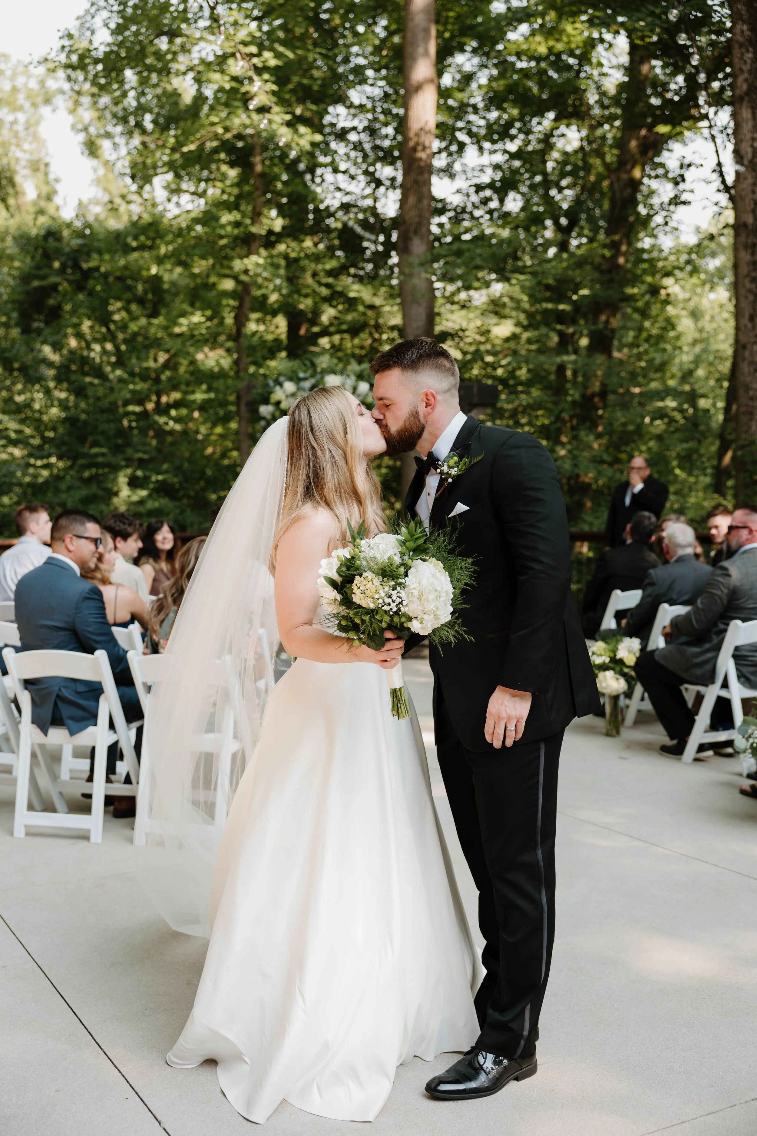 A bride and groom kiss during their outdoor wedding ceremony surrounded by trees, with guests seated on white chairs in the background.