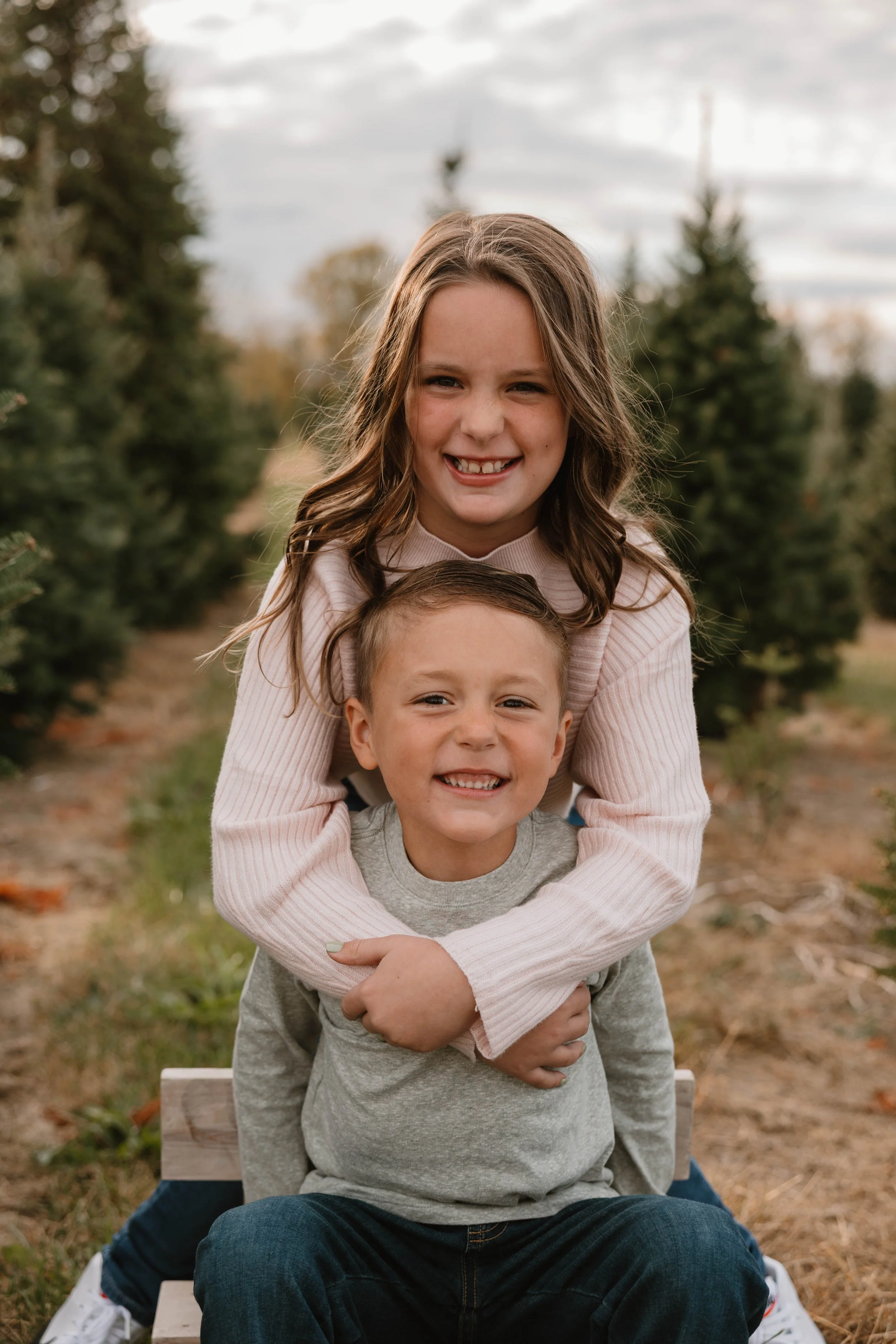 A girl and a boy smiling, sitting outside among evergreen trees.